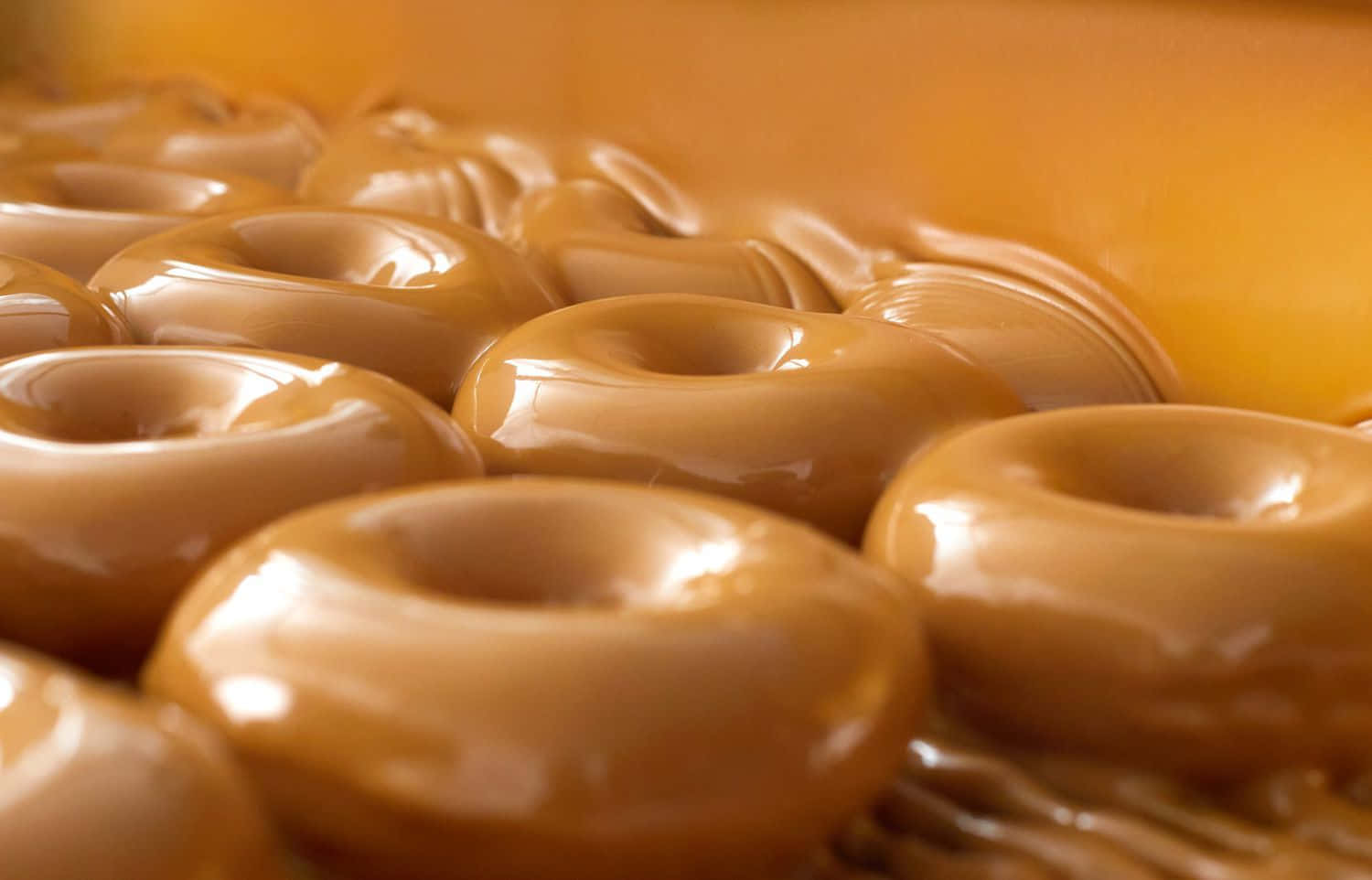 Glazed Donut 桌布 壁紙 — 特寫鏡頭：閃亮焦糖釉面冬甩，排列成溫暖金色調，配以柔和散景背景。
