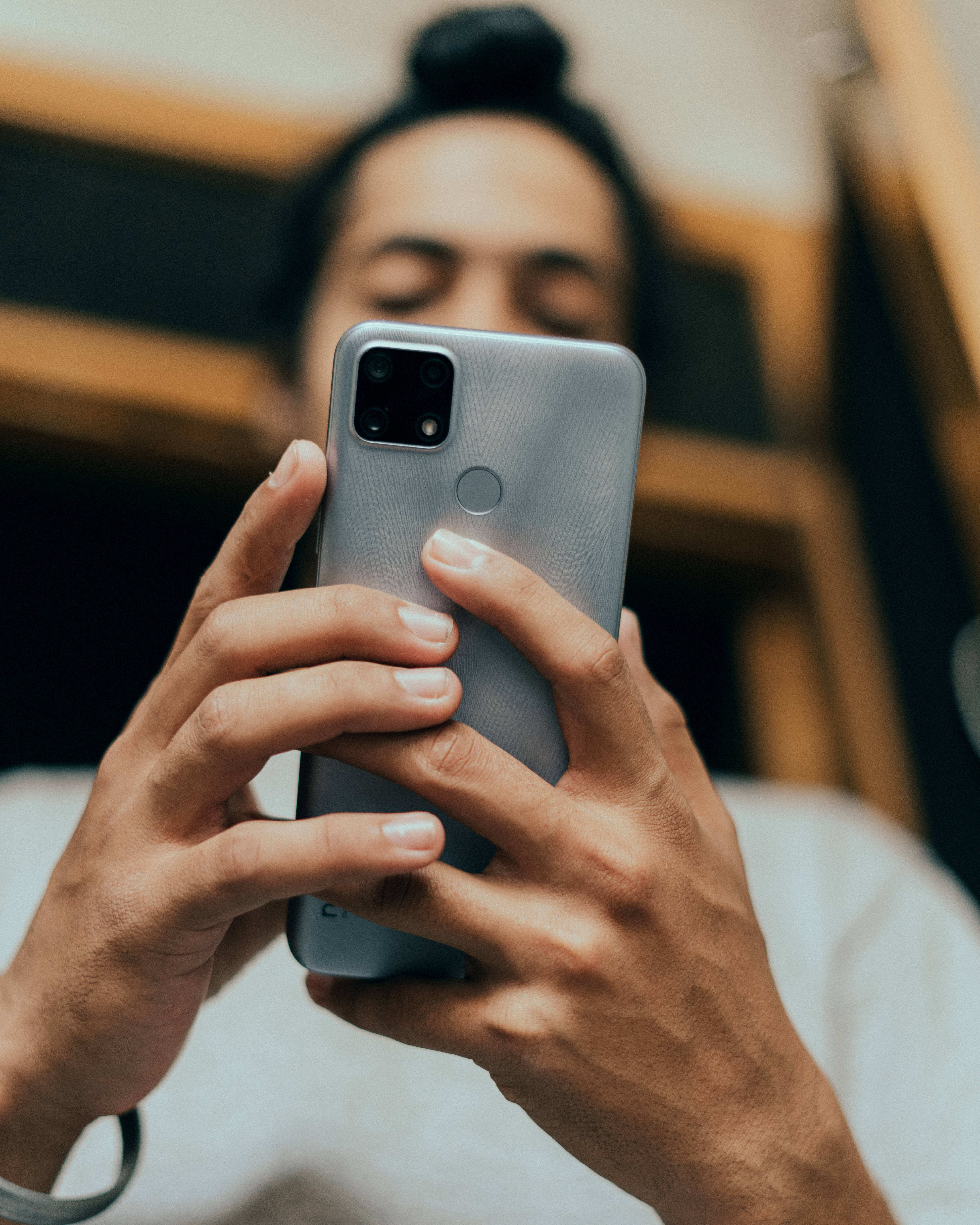 Realme Narzo 20 壁紙 — 特寫鏡頭，雙手握著一部銀色四鏡頭智能手機，背景模糊中有一個人閉著眼睛看著。
