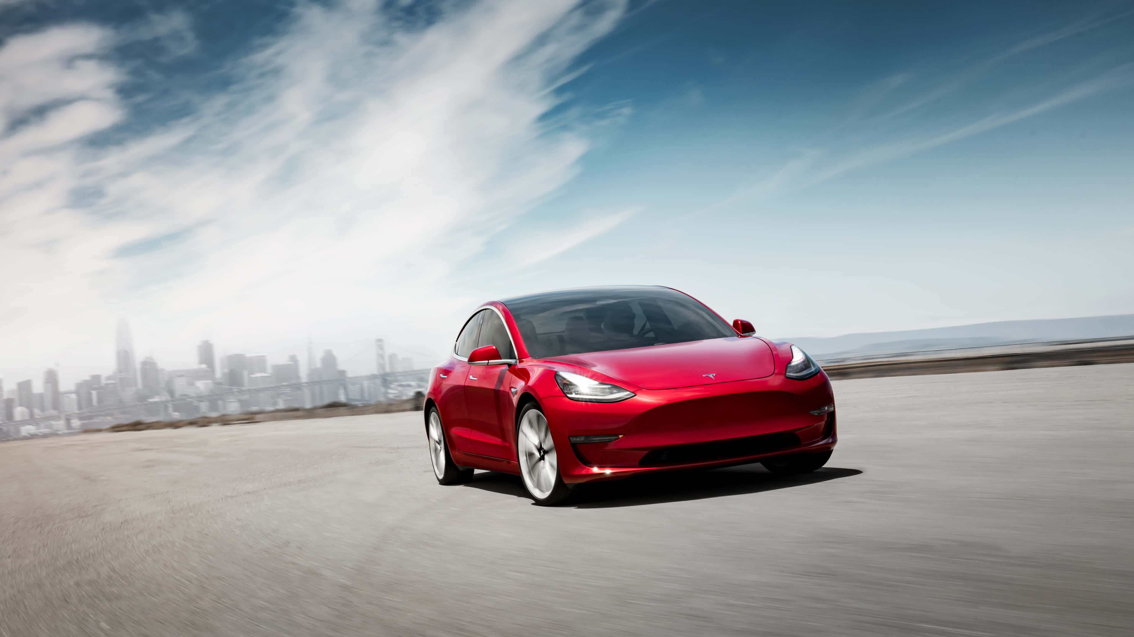 特斯拉 Model 3 的工藝之美 壁紙 — 一輛鮮紅色的Tesla Model 3在部分多雲的藍天下，沿著一條鋪砌的道路疾馳，背景是朦朧的城市天際線。