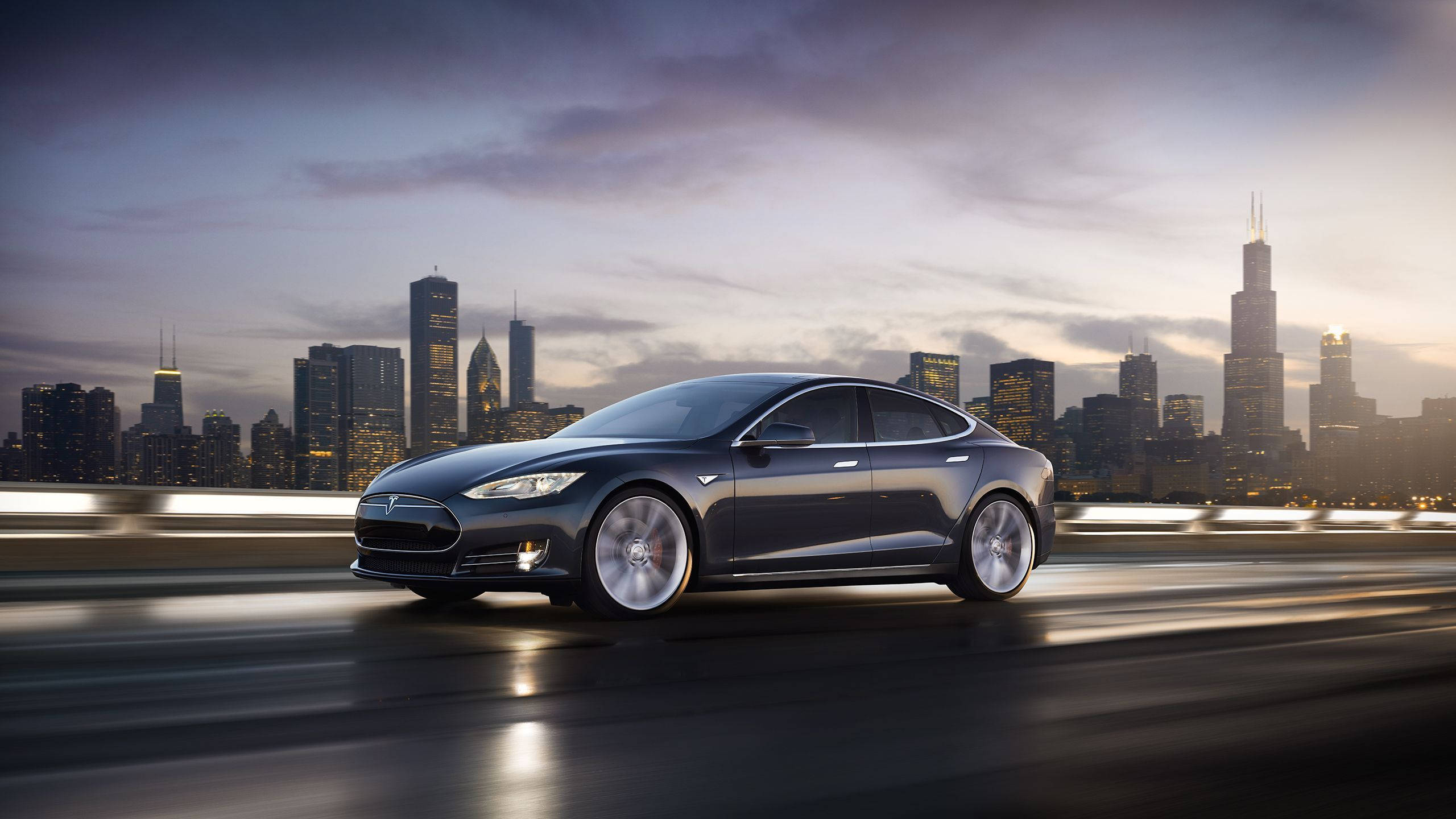 特斯拉 Model S 城市風景 壁紙 — 一架時尚的深灰色Tesla Model S在高速公路上巡航，芝加哥璀璨的天際線在黃昏時分在它背後閃耀。