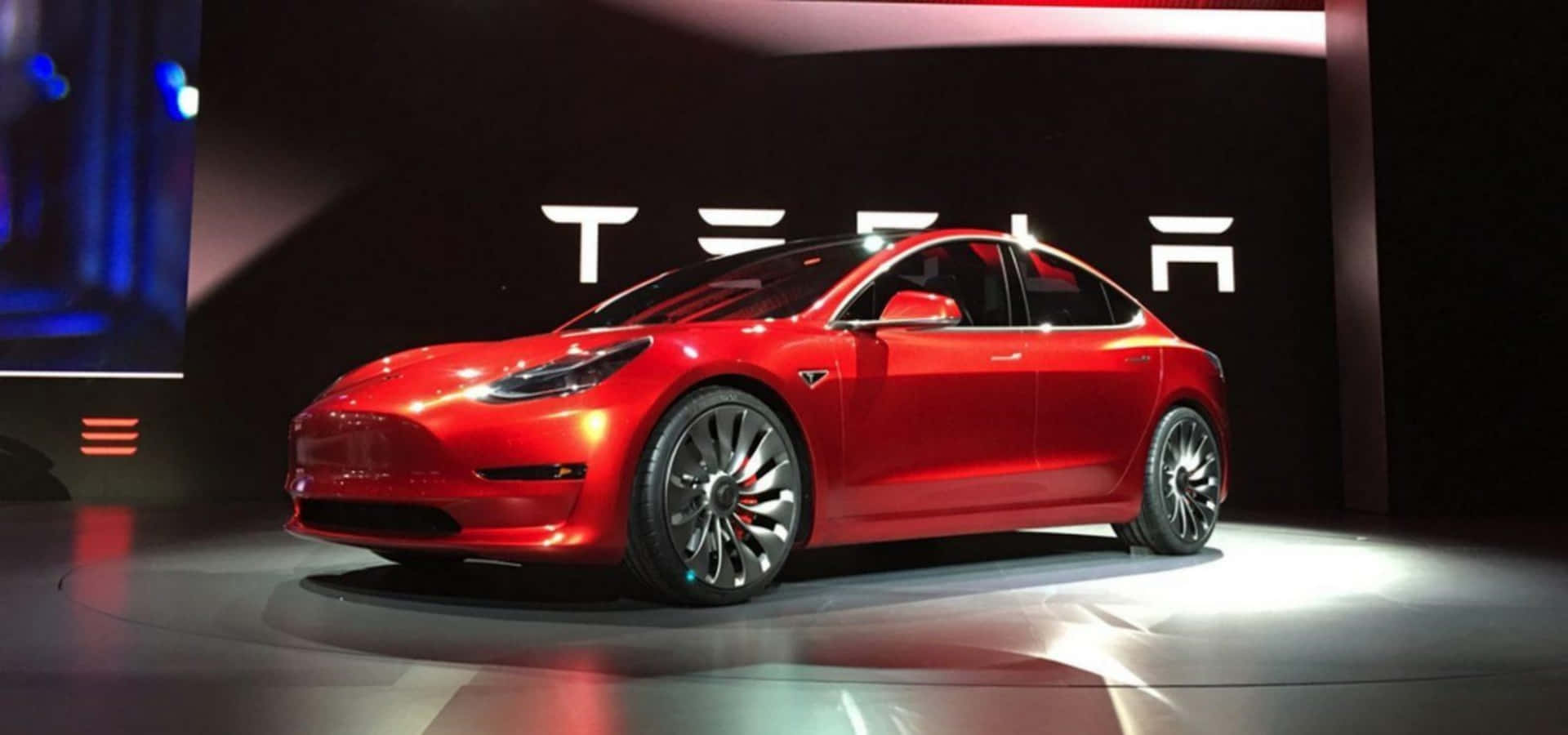 特斯拉Model 3 — 超越期待的創新之作 壁紙 — 一輛光澤的紅色 Tesla Model 3 房車在明亮的聚光燈下於圓形舞台上展示，背後 Tesla 標誌閃閃發光。