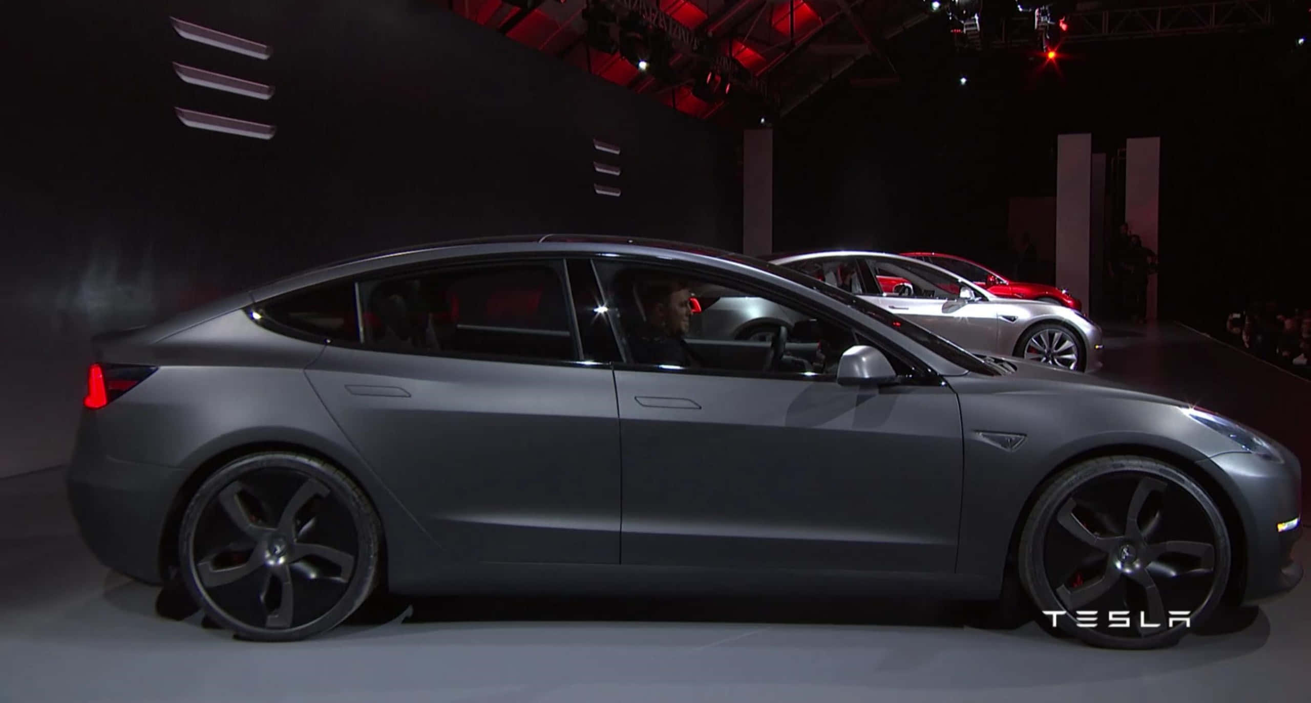 特斯拉Model 3：電動車的未來 壁紙 — 一輛時尚的啞光灰色Tesla Model 3在昏暗的舞台上展示，配有紅色重點照明，背景中可見另一輛車。
