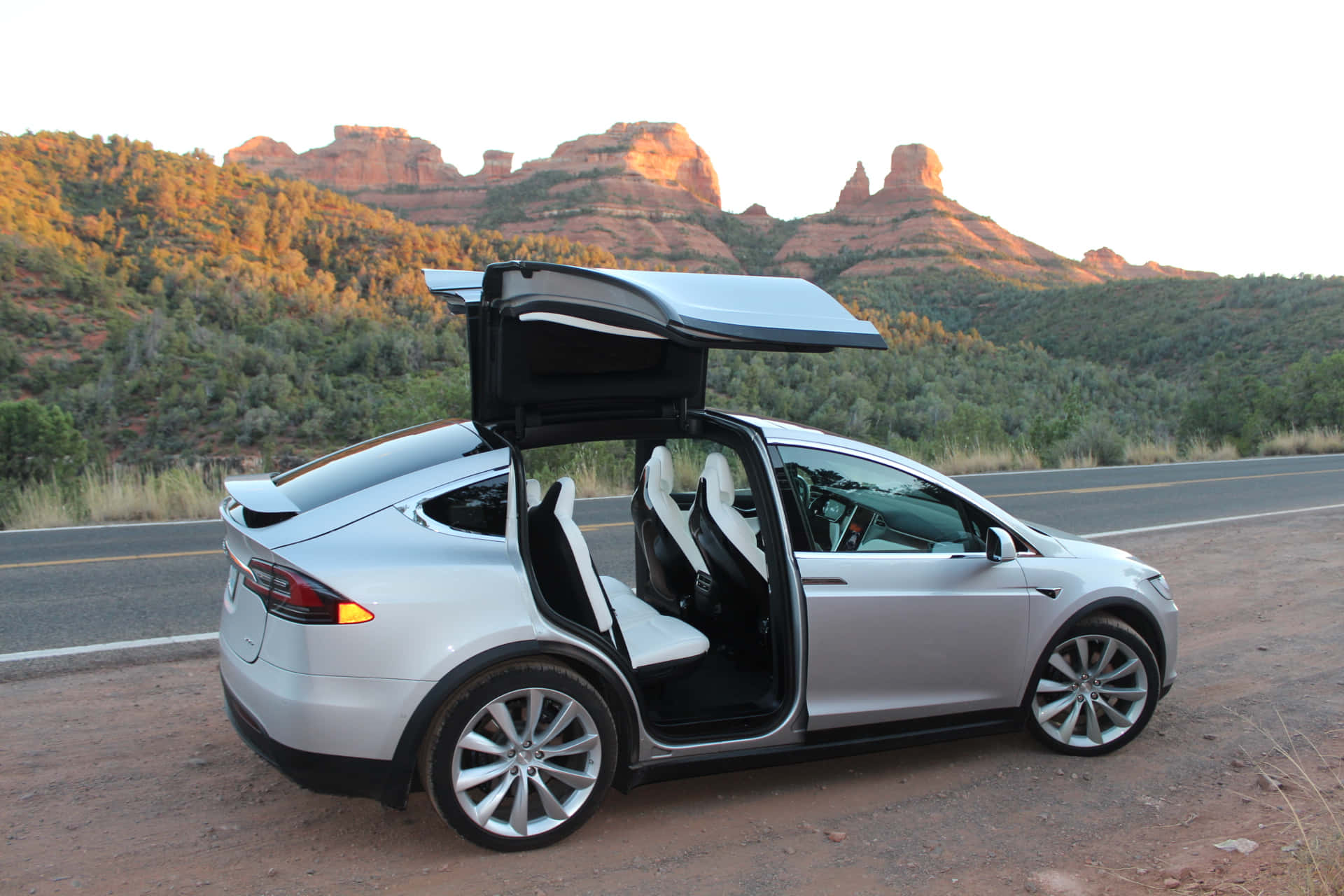銀色 Tesla Model X 打開車門，停在日落時分紅色岩層附近的沙漠公路上。