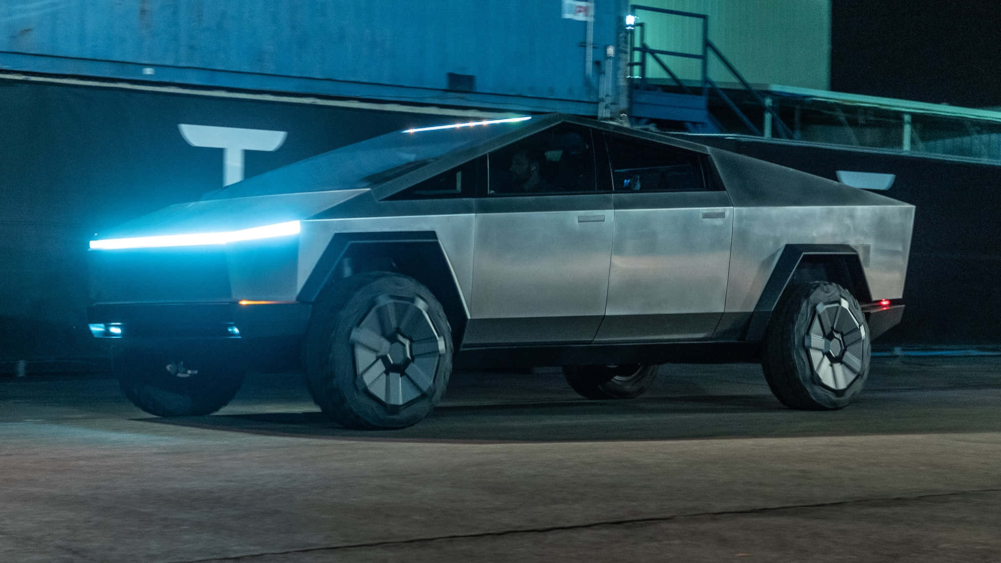 特斯拉未來感十足的Cybertruck即將上路 壁紙 — 一輛未來感十足的銀色 Tesla Cybertruck 在夜間昏暗的工業車庫中，在霓虹藍色頭燈下閃閃發光。