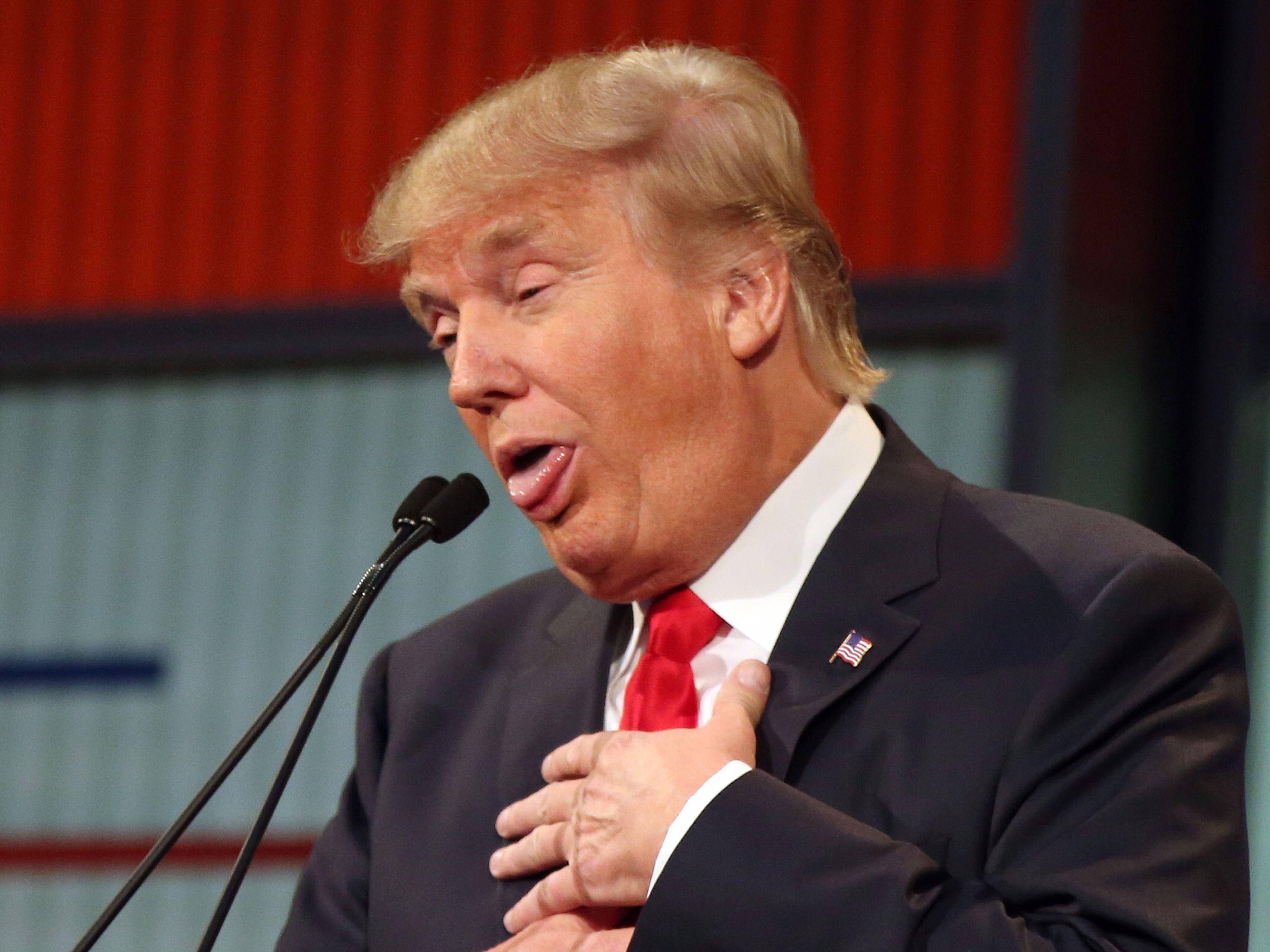 特朗普吐舌微笑 壁紙 — 一名身穿深色西裝和紅色領帶的男子，在紅色和藍色背景下，將手放在心口上，熱情地對著麥克風說話。