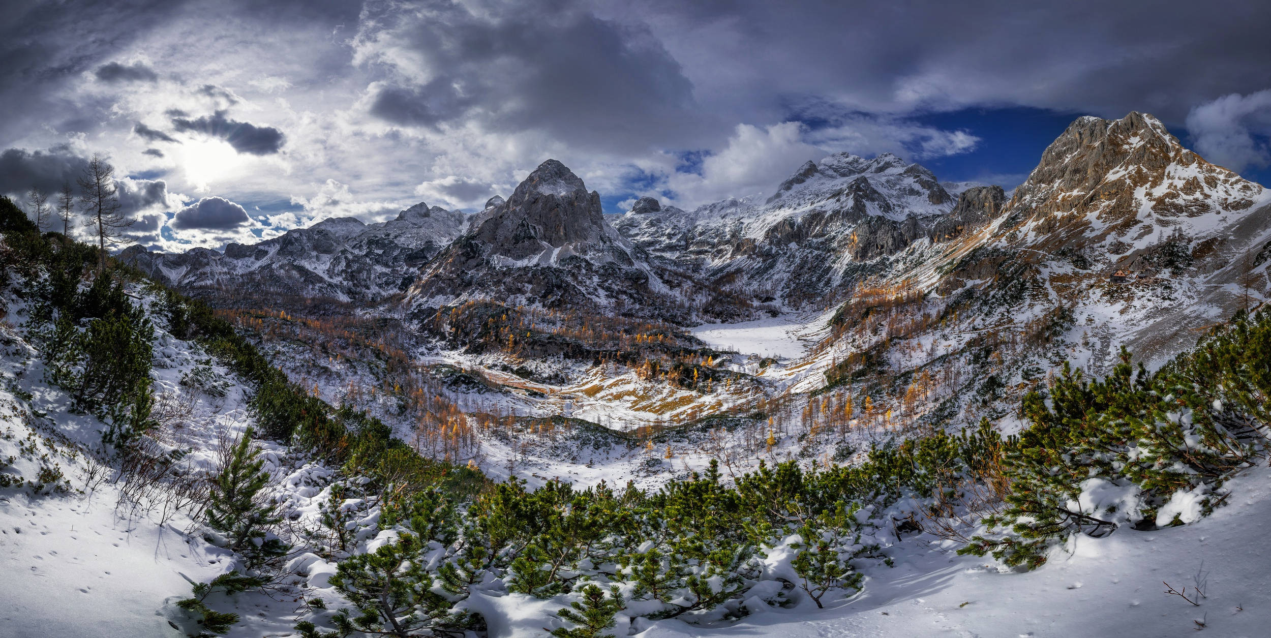特里格拉夫山峰雪景 壁紙 — 白雪覆蓋著崎嶇的山峰，在戲劇性的多雲天空下，山谷底部有藍色斑塊和金色光芒。