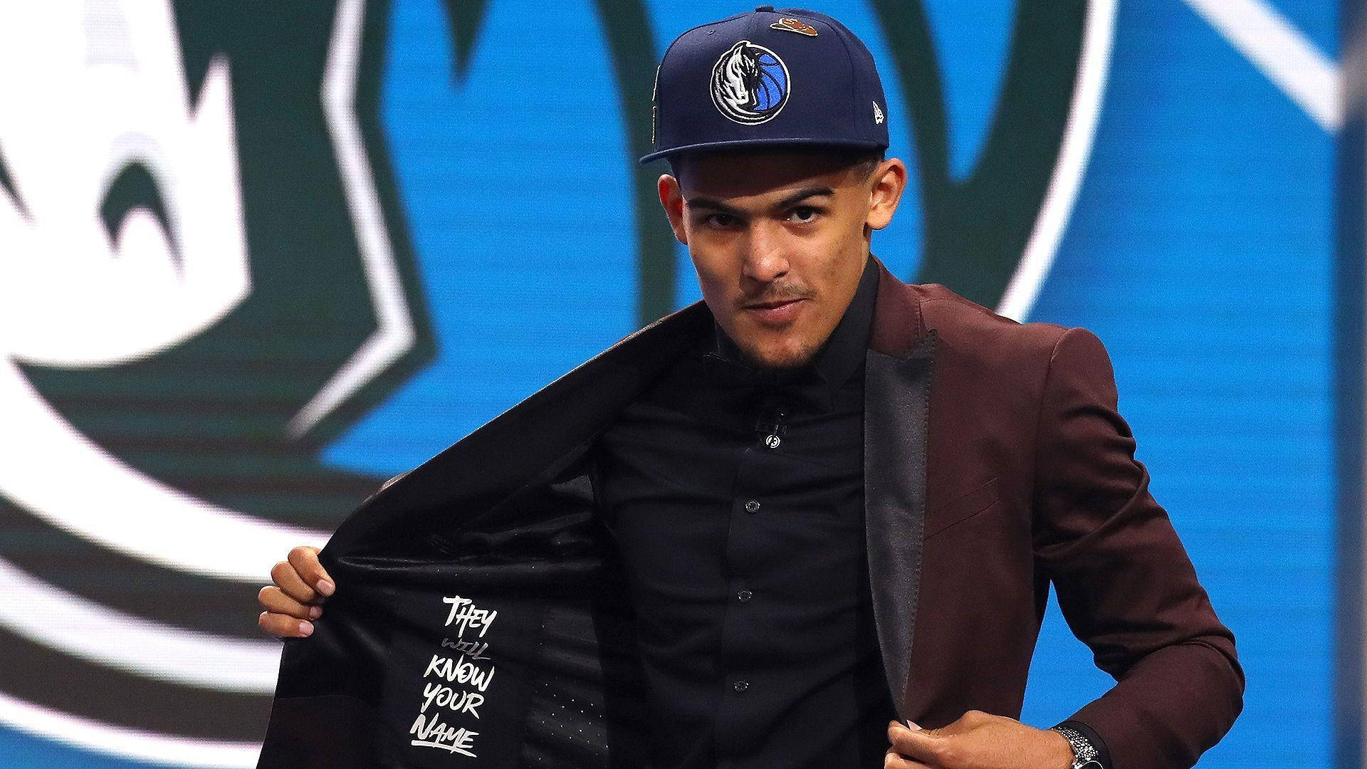 特雷·楊 2018 NBA 新秀選秀 壁紙 — 一位身穿酒紅色西裝外套和達拉斯獨行俠帽子的籃球運動員，戲劇性地揭開一件寫有「They Know Your Name」的外套。