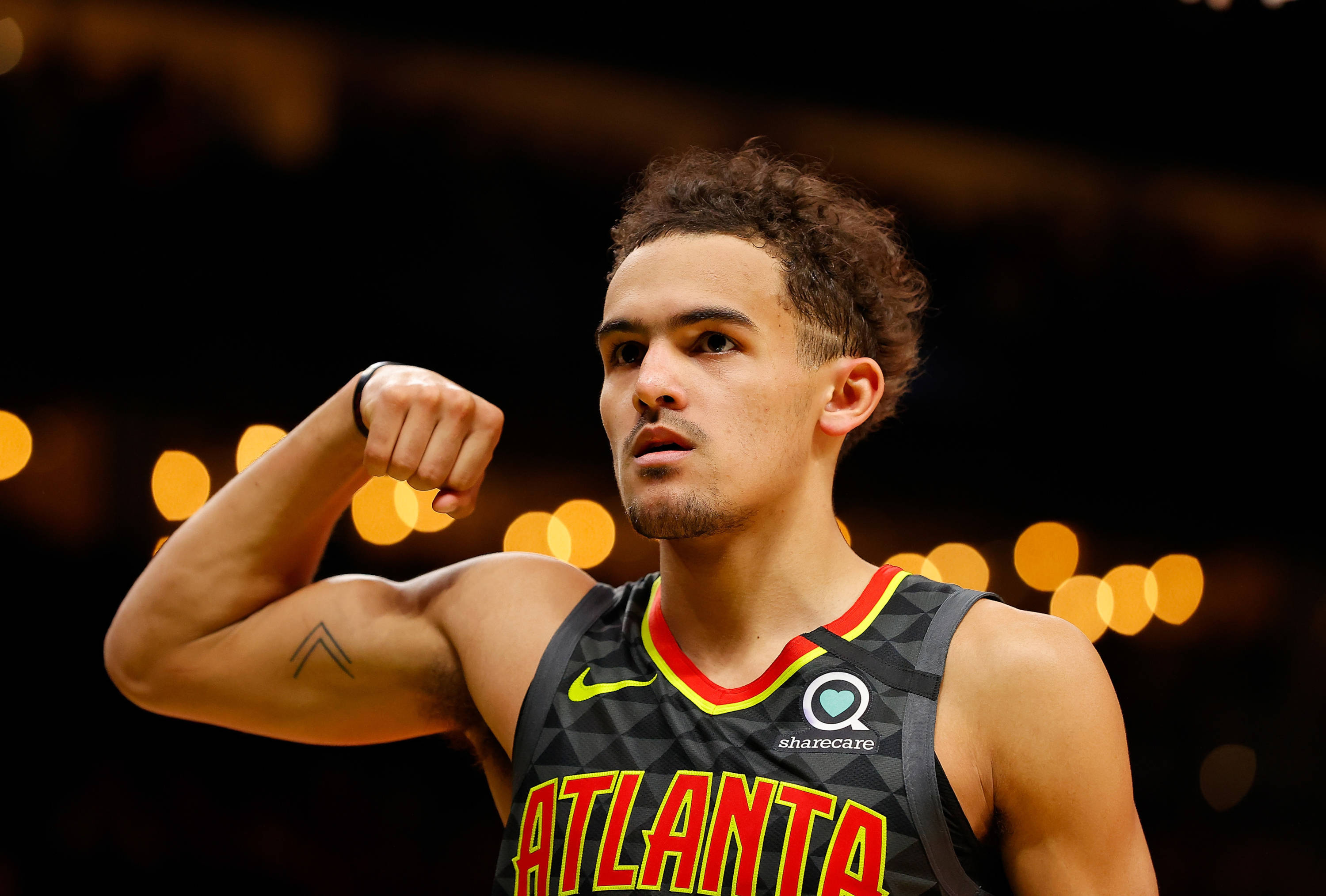 特雷·楊NBA亞特蘭大球員 壁紙 — 一個專注的籃球運動員在場館燈光下彎曲他的二頭肌，背景有模糊的金色散景。