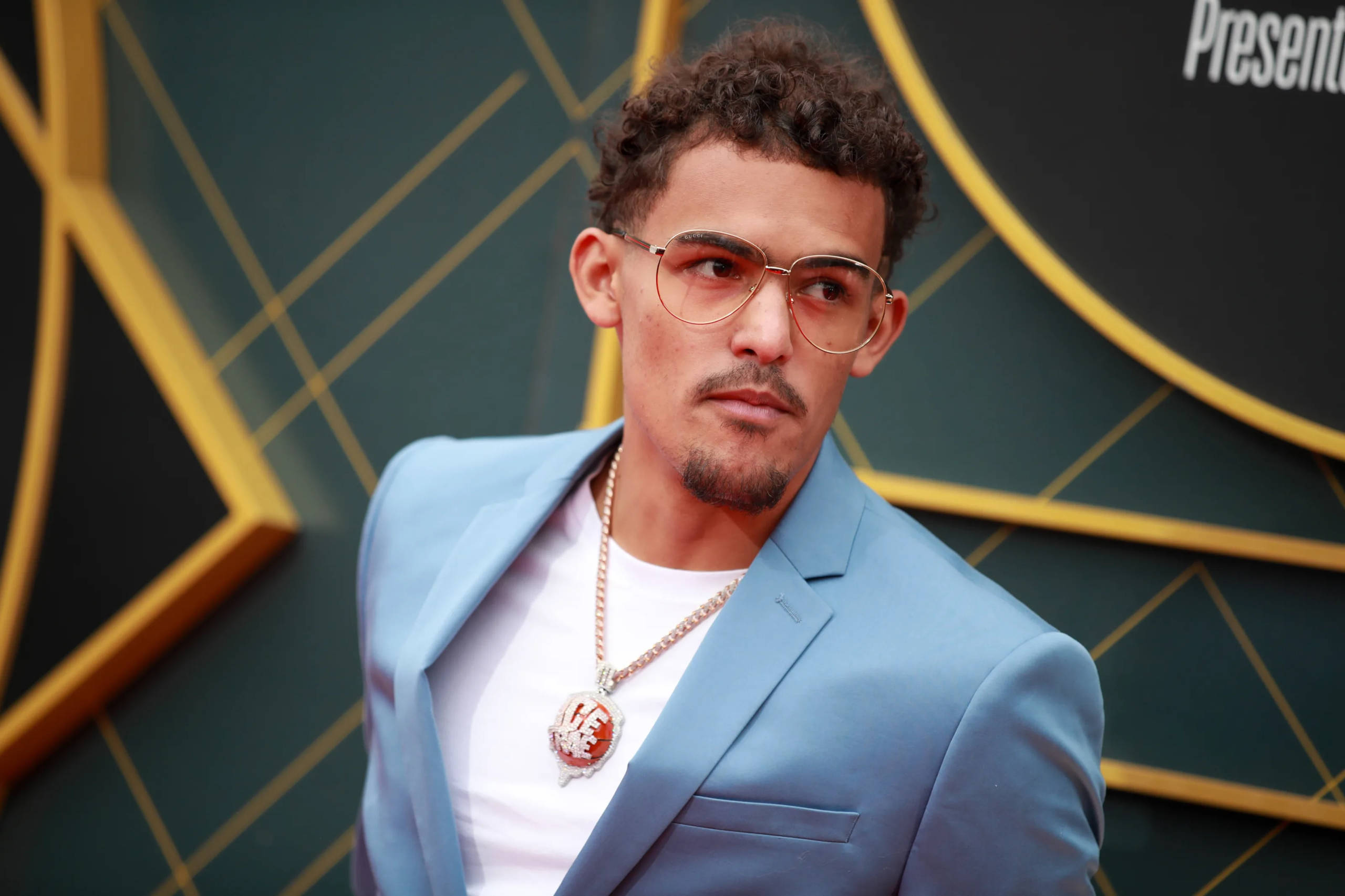 特雷·楊NBA頒獎典禮 壁紙 — 一位時尚男士身穿淺藍色西裝外套，戴著金邊眼鏡，在幾何金色和深綠色背景前自信地擺姿勢。