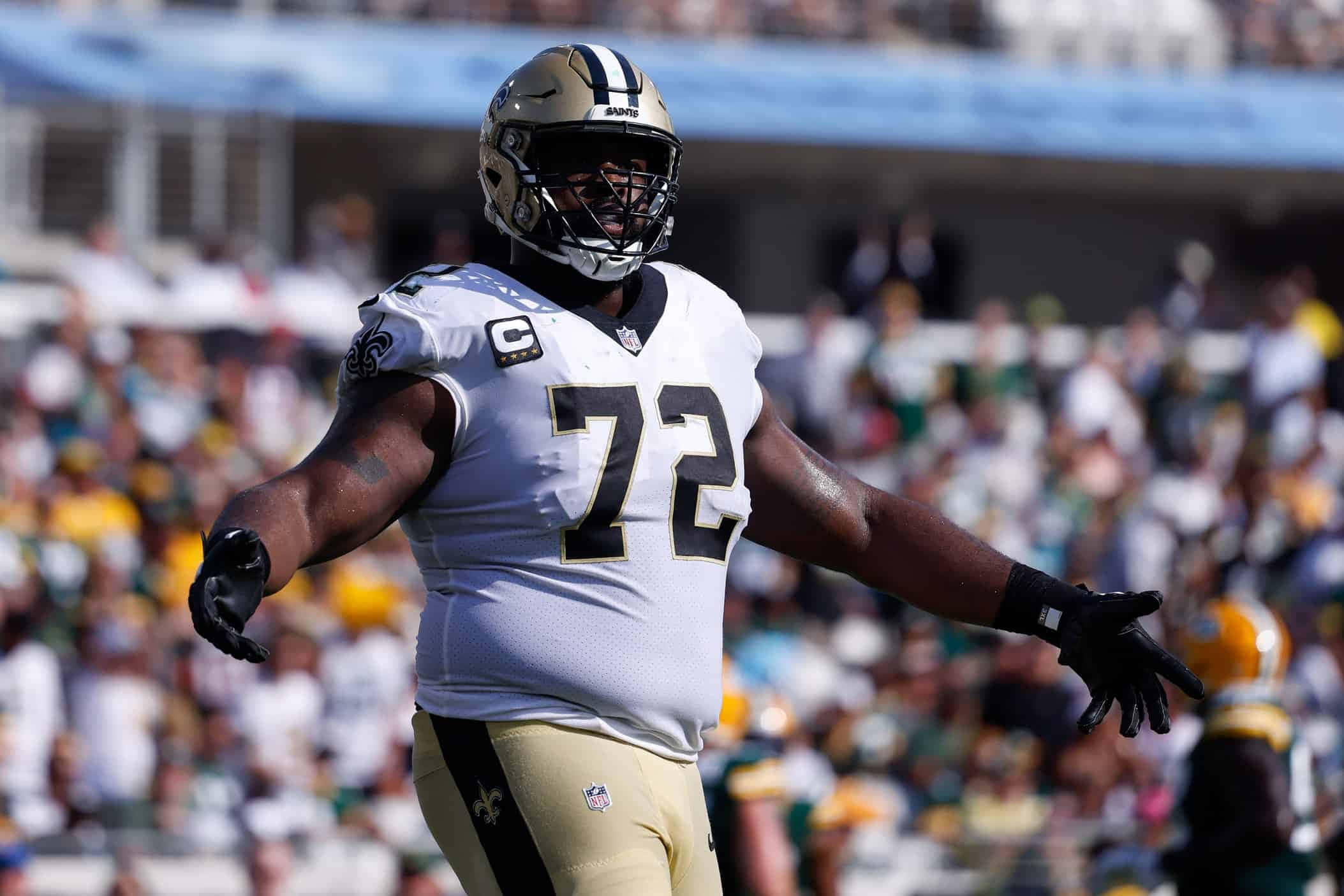 特龍·阿姆斯特德 新奧爾良聖徒隊 NFL Tackle 壁紙 — 一位身穿白色和金色球衣的新奧爾良聖徒隊球員在陽光明媚的比賽日場地上張開雙臂慶祝。