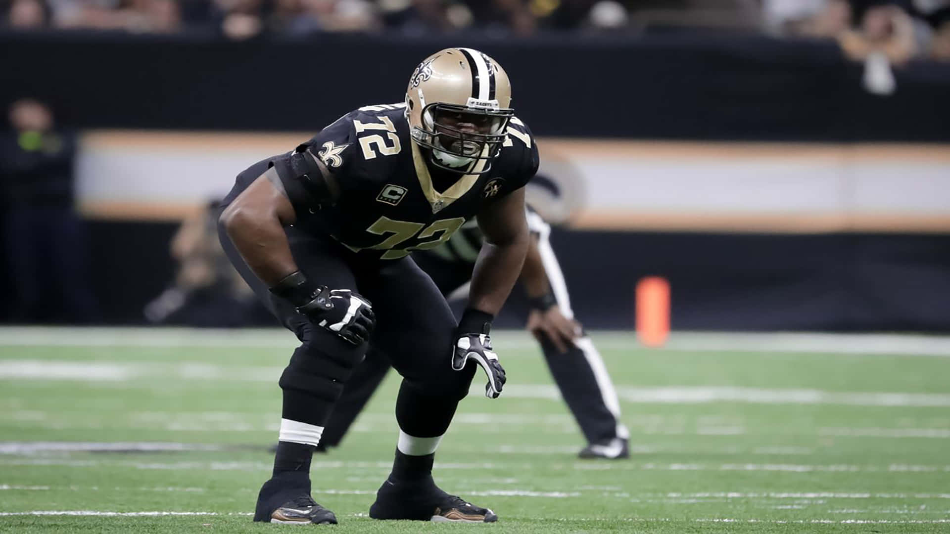 特龍·阿姆斯特德 紐奧良聖徒隊 NFL球員 壁紙 — 一名新奧爾良聖徒隊的美式足球員穿著黑色和金色裝備在比賽中蹲低在場上。