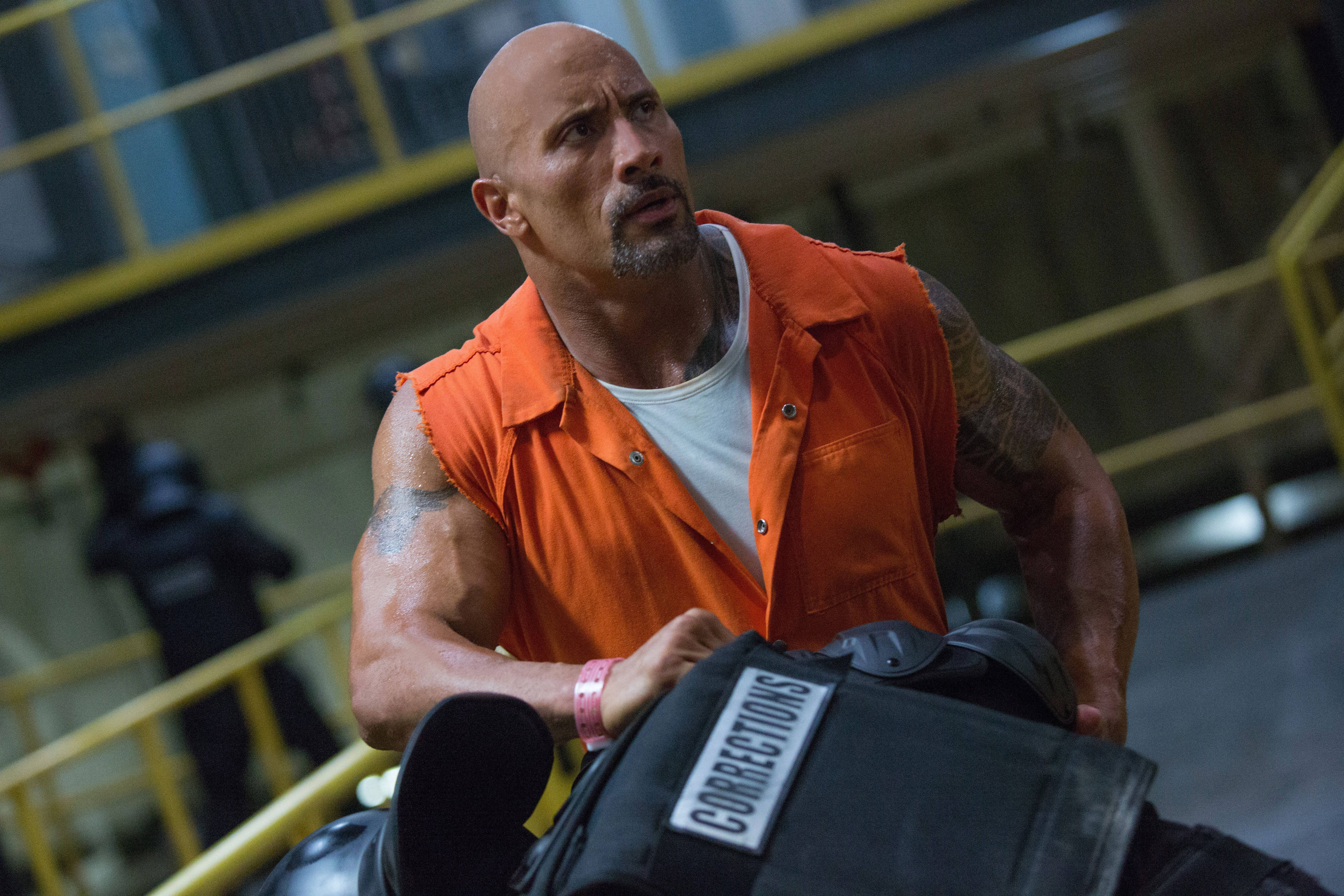 The Rock 桌布 壁紙 — 狄維·莊遜身穿橙色監獄連身衣，在陰暗的工業設施中用力推動沉重的黑色矯正推車。