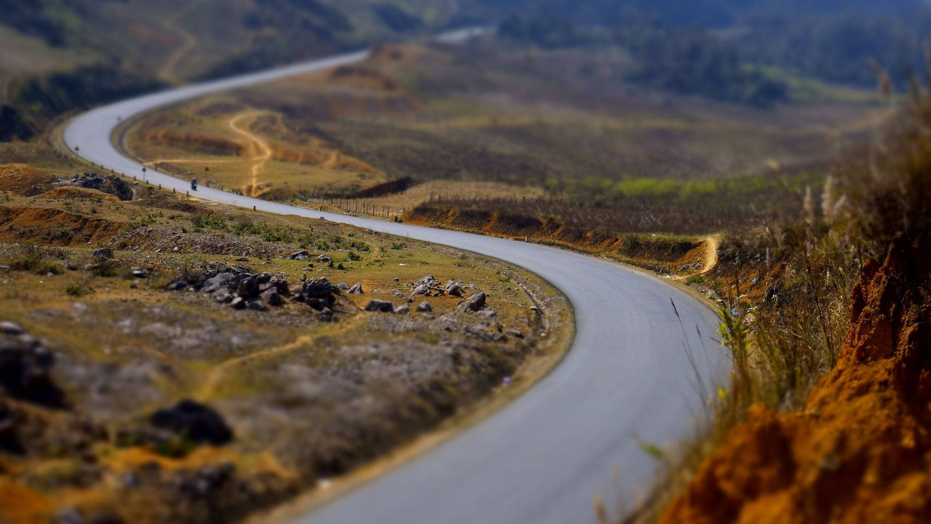 狹窄彎曲道路之旅 壁紙 — 一條蜿蜒的瀝青道路穿過崎嶇的丘陵地帶，有乾草和散落的岩石，在柔和的光線下。