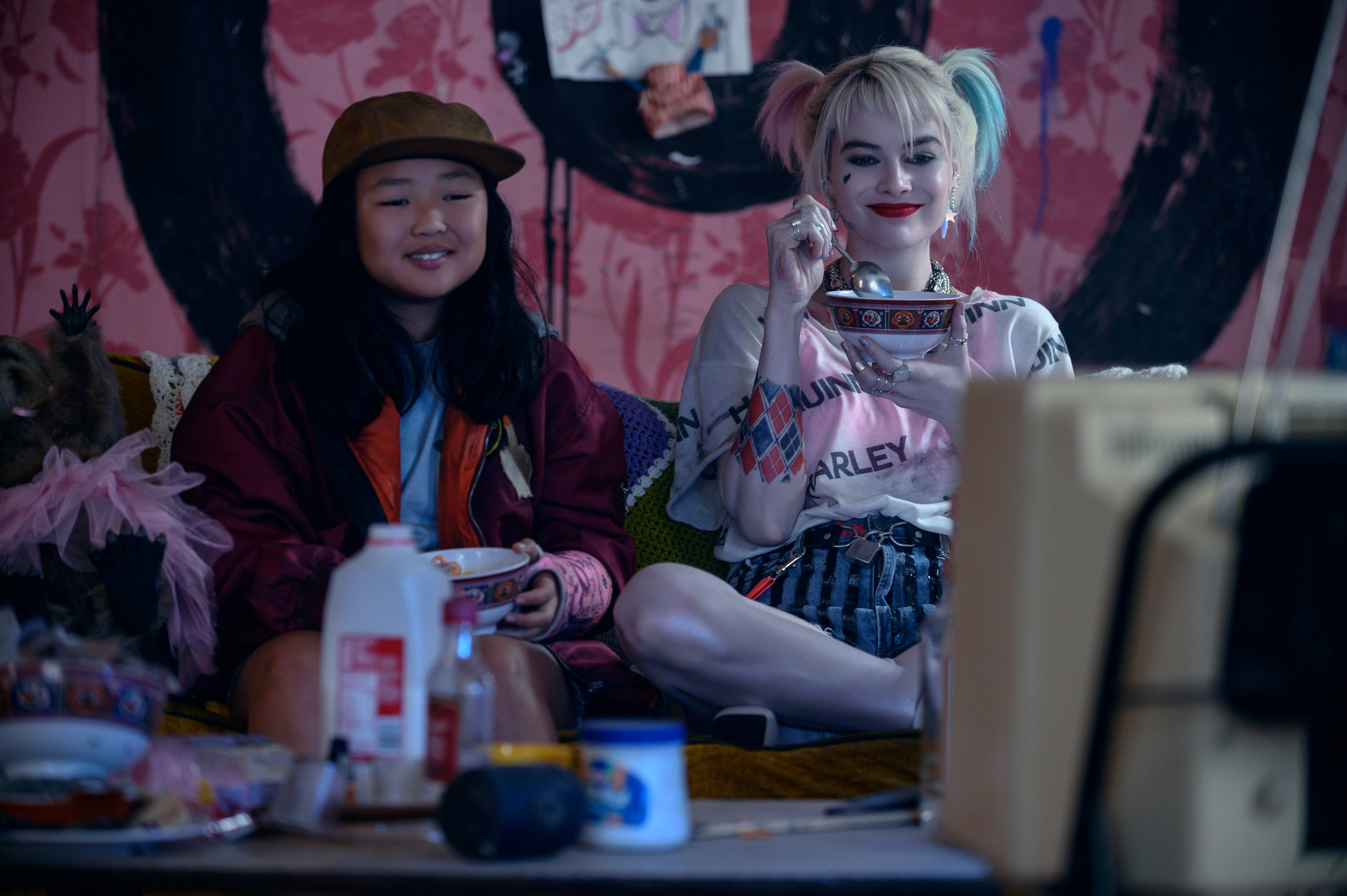 《猛禽小隊》哈莉·奎茵與卡珊德拉 壁紙 — 兩個女人坐在雜亂房間的沙發上，一個紮著粉藍色辮子正在從碗裡吃東西，另一個在她旁邊微笑。