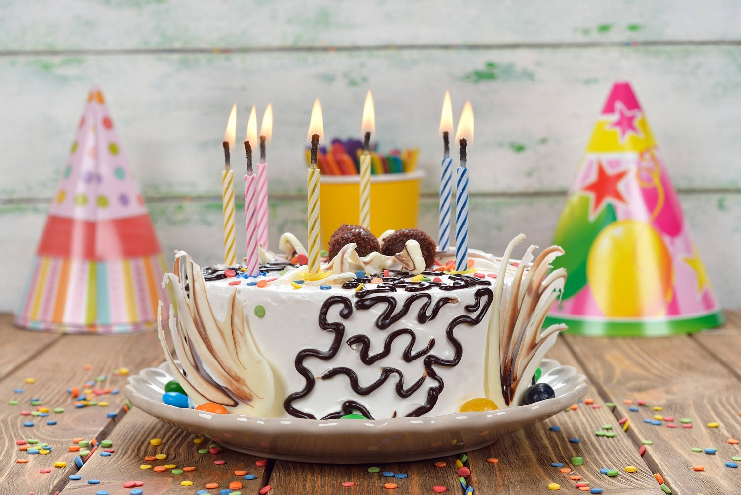 獨特裝飾生日蛋糕蠟燭 壁紙 — 一個節日生日蛋糕，上面有燃燒的蠟燭、朱古力醬和彩色糖粒，放在一個木桌上，周圍有派對帽和五彩紙屑。