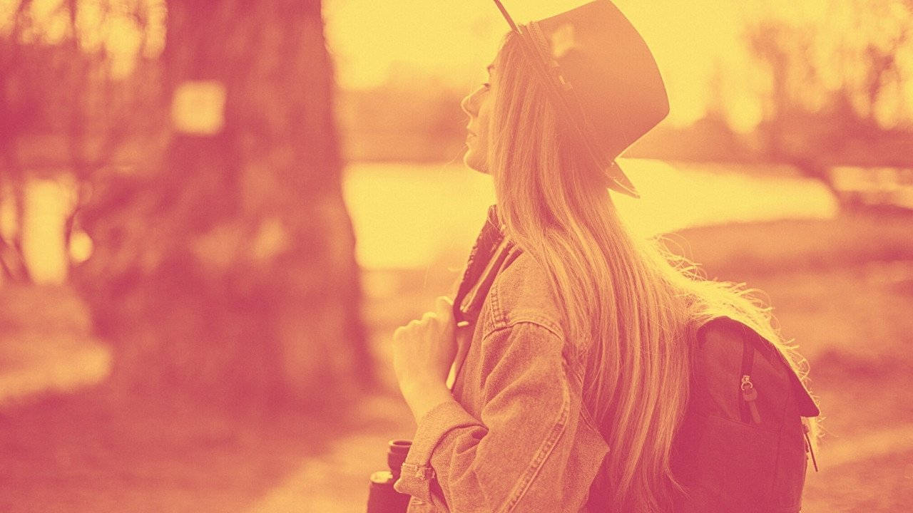 獨立旅行者 壁紙 — 一位長髮年輕女性戴著帽子和背包，在溫暖的金色陽光下若有所思地凝視遠方。