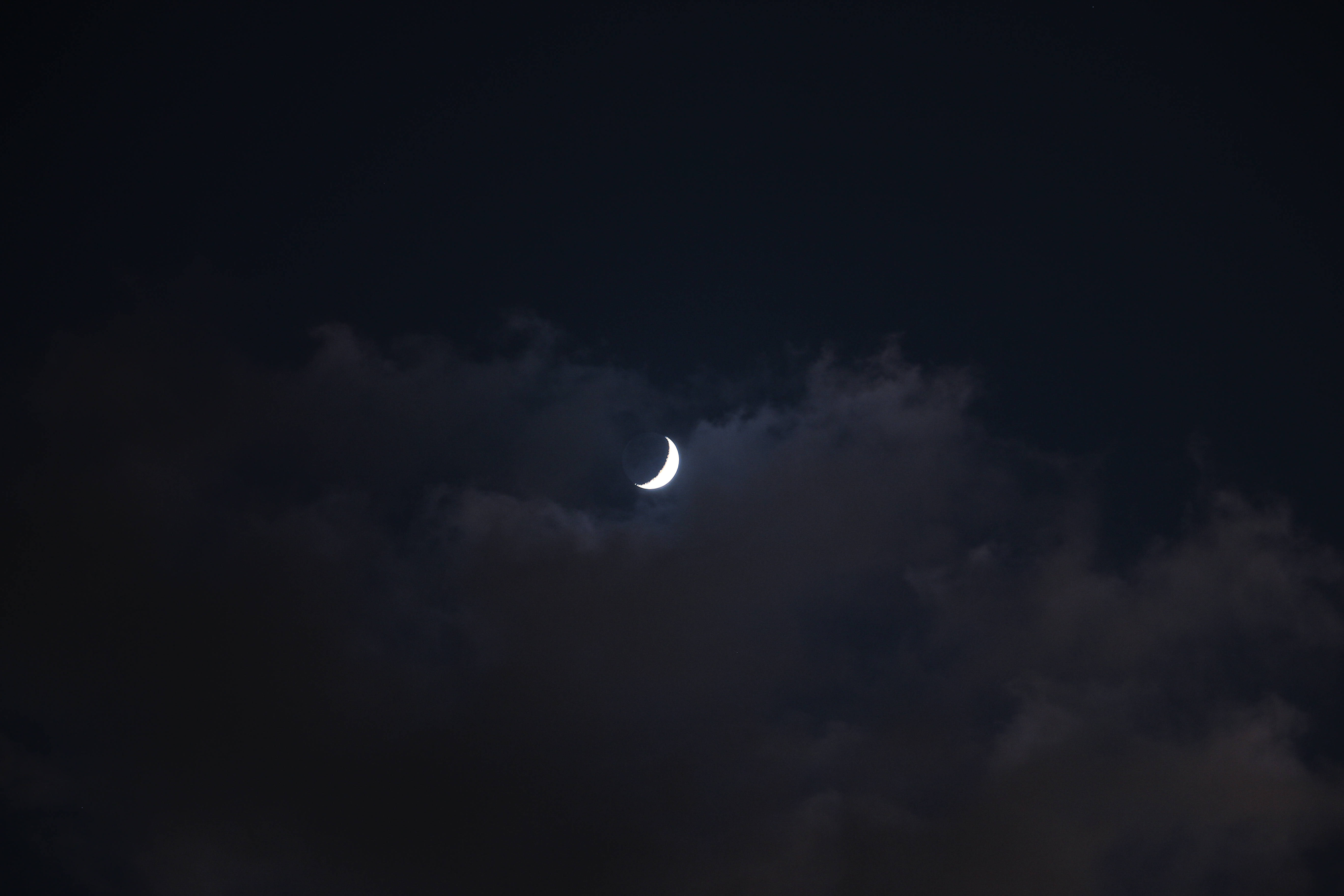獨自升起的美學月 壁紙 — 一彎纖細的月牙在深邃的夜空中，透過飄動的烏雲柔和地發光。