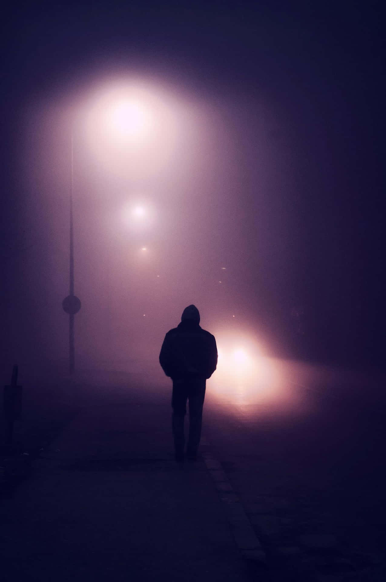 在發光的燈光下，夜間霧氣瀰漫的城市街道上獨自行走的人的剪影。