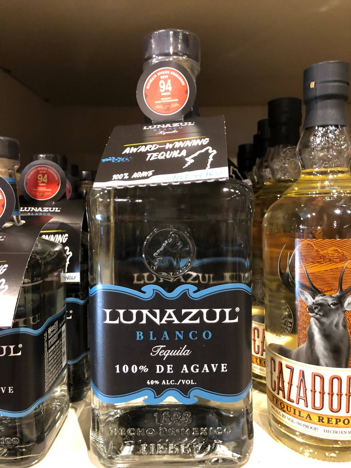 獲獎殊榮Lunazul Blanco龍舌蘭酒 壁紙 — 一個透明嘅Lunazul Blanco龍舌蘭酒瓶，黑色標籤同藍色點綴，擺喺貨架上，旁邊有其他酒瓶。