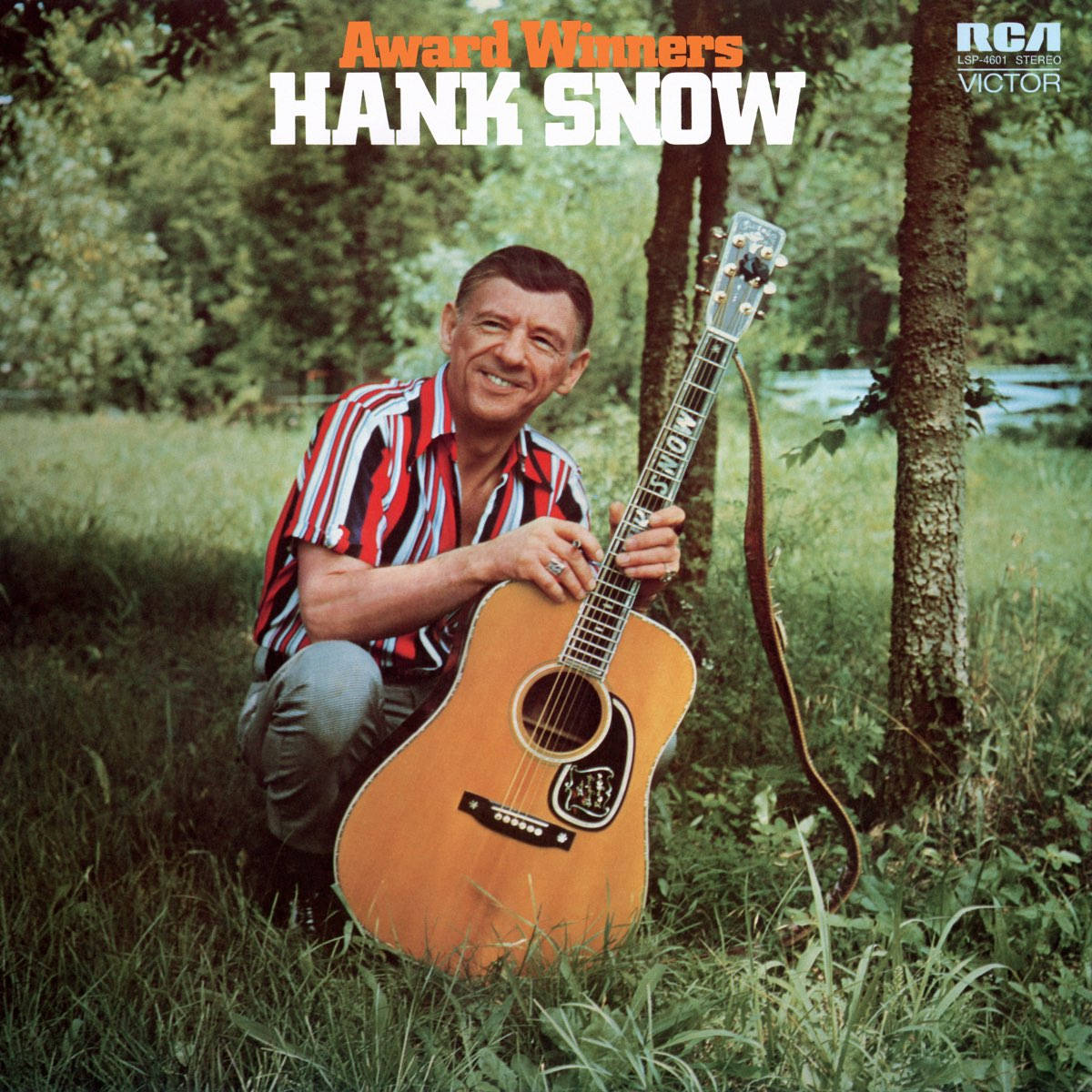 獲獎藝術家漢克·斯諾肖像 壁紙 — Hank Snow 在陽光明媚的戶外環境中，跪在草地上，手持木結他，在樹旁露出溫暖的笑容。