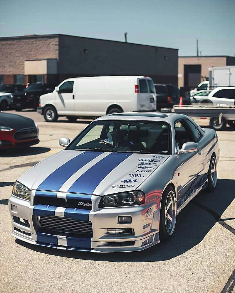 銀色日產Skyline R34，帶有藍色賽車條紋，停泊在陽光明媚的停車場。