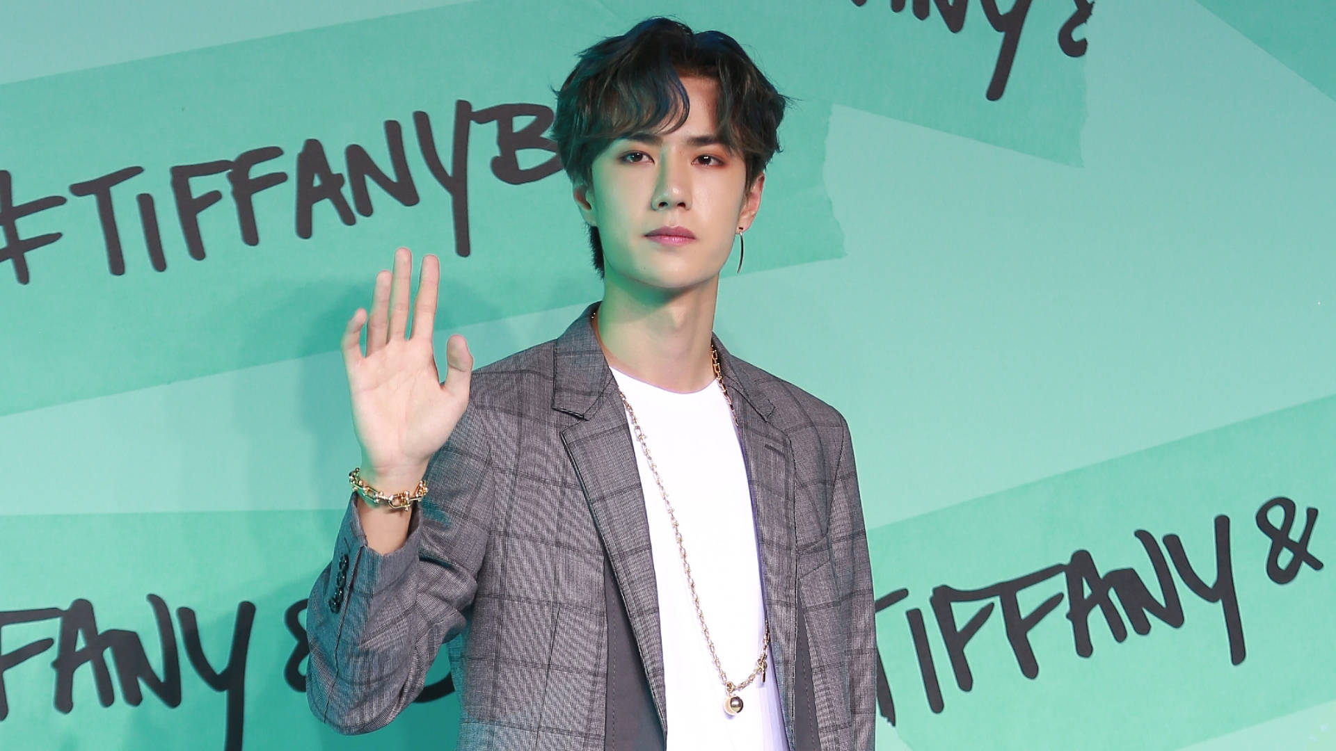 王一博 x Tiffany & Co. 壁紙 — 一位身穿格仔西裝外套的年輕男士，在Tiffany & Co的藍綠色背景前向鏡頭揮手。