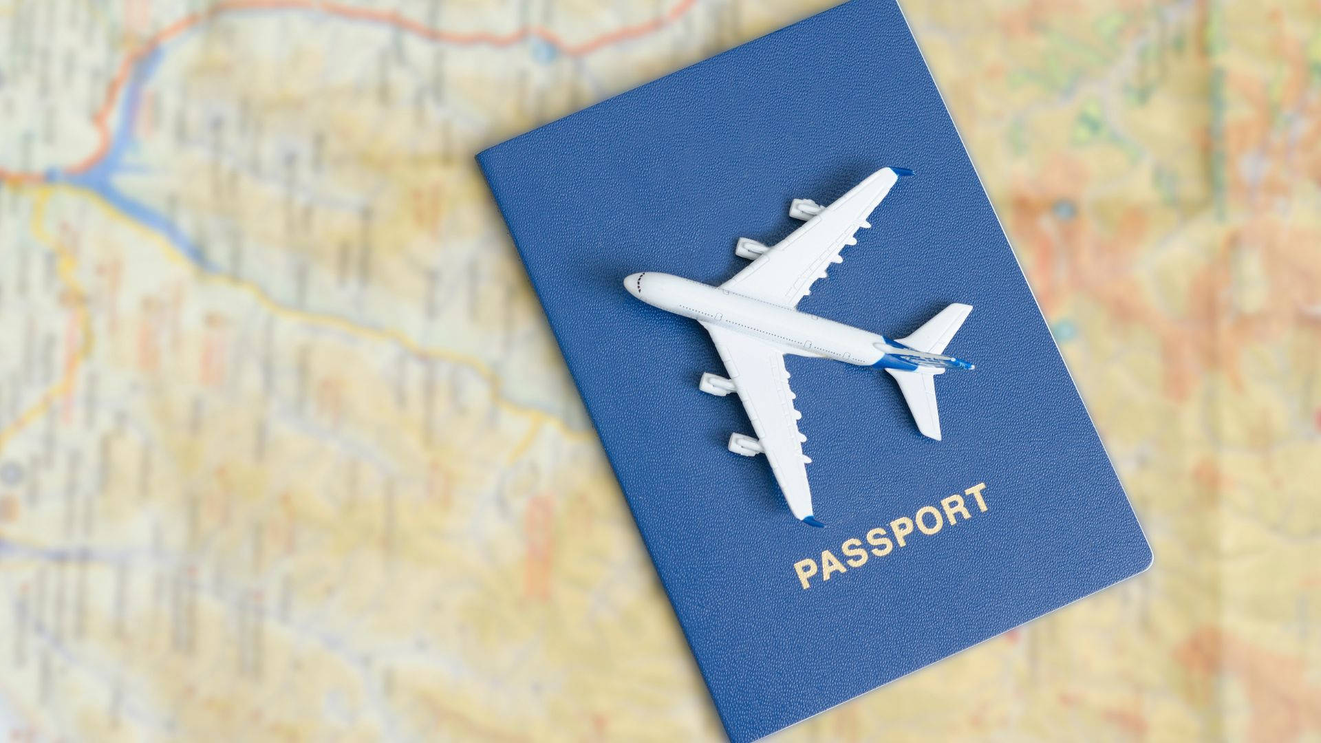 玩具飛機與護照俯瞰旅行地圖 壁紙 — 一架白色飛機模型放在一本藍色護照上，護照則放在一張詳細的地圖上，暗示全球旅行。