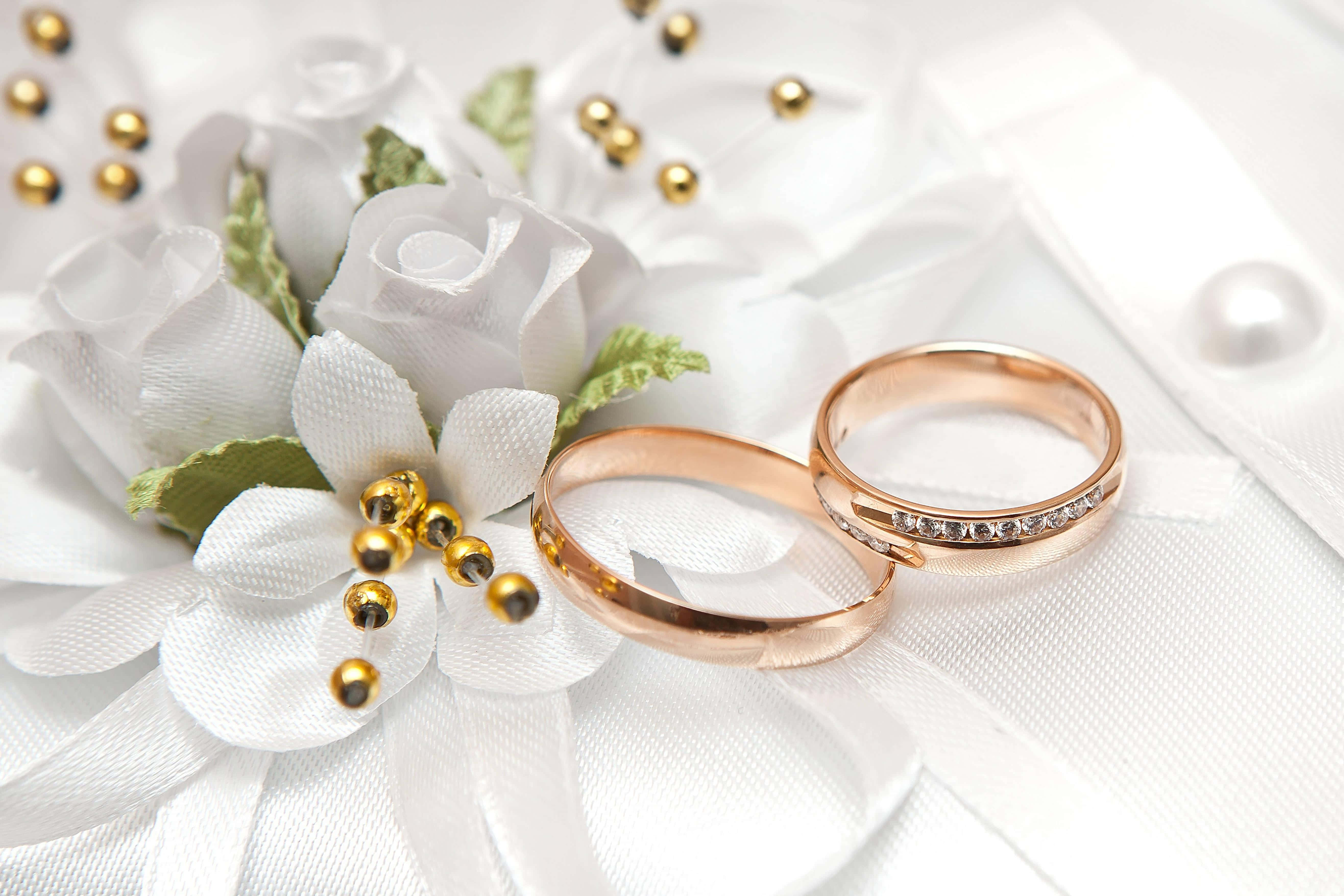 玫瑰金戒指圖像 壁紙 — 兩枚玫瑰金結婚戒指放在白色布藝玫瑰和金珠旁邊，置於柔軟的白色緞面之上。
