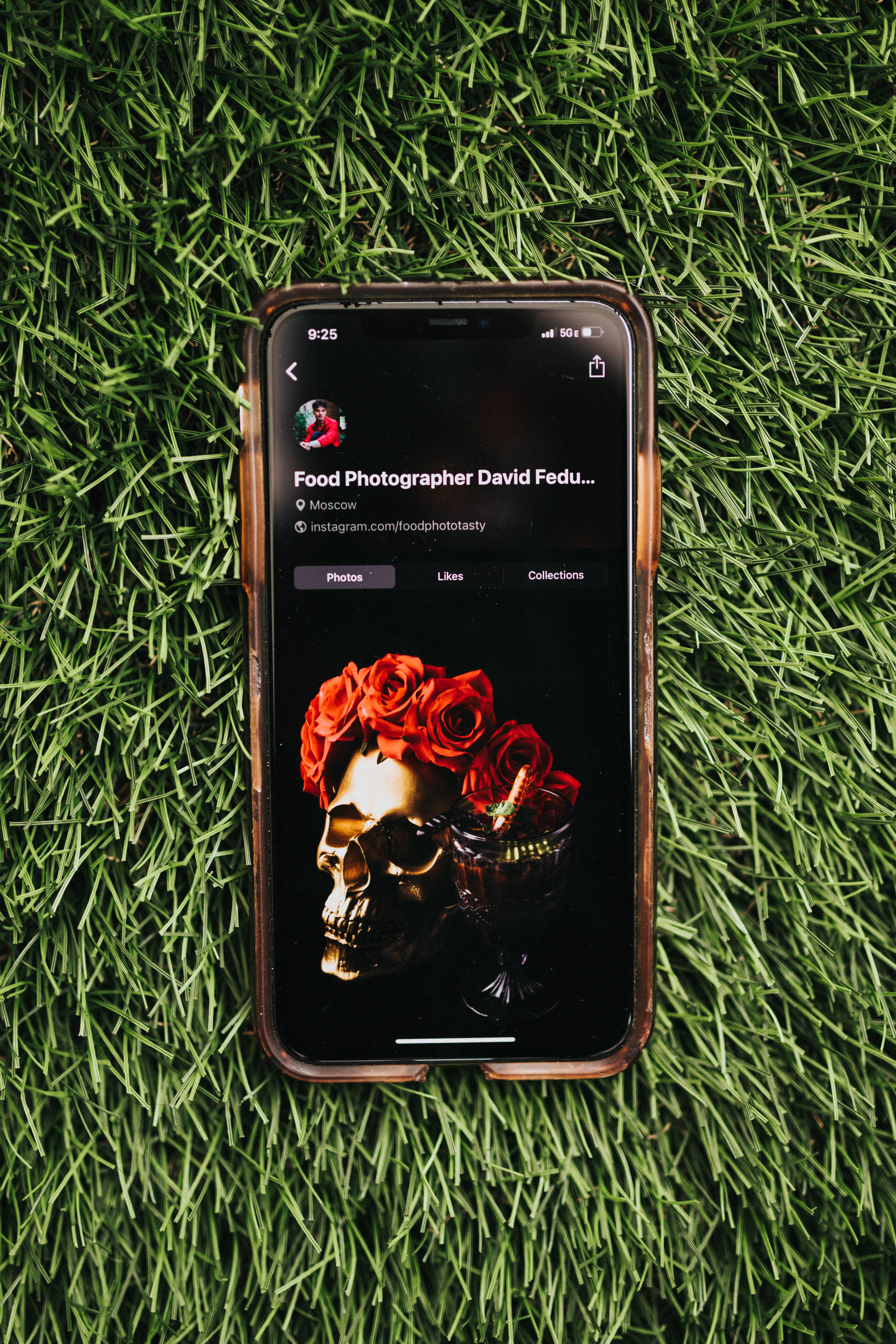 玫瑰骷髏iPhone iOS 壁紙 — 一部顯示美食攝影師Instagram個人資料的智能手機放在鮮綠色草地上，展示一個飾有紅玫瑰的金色頭骨。
