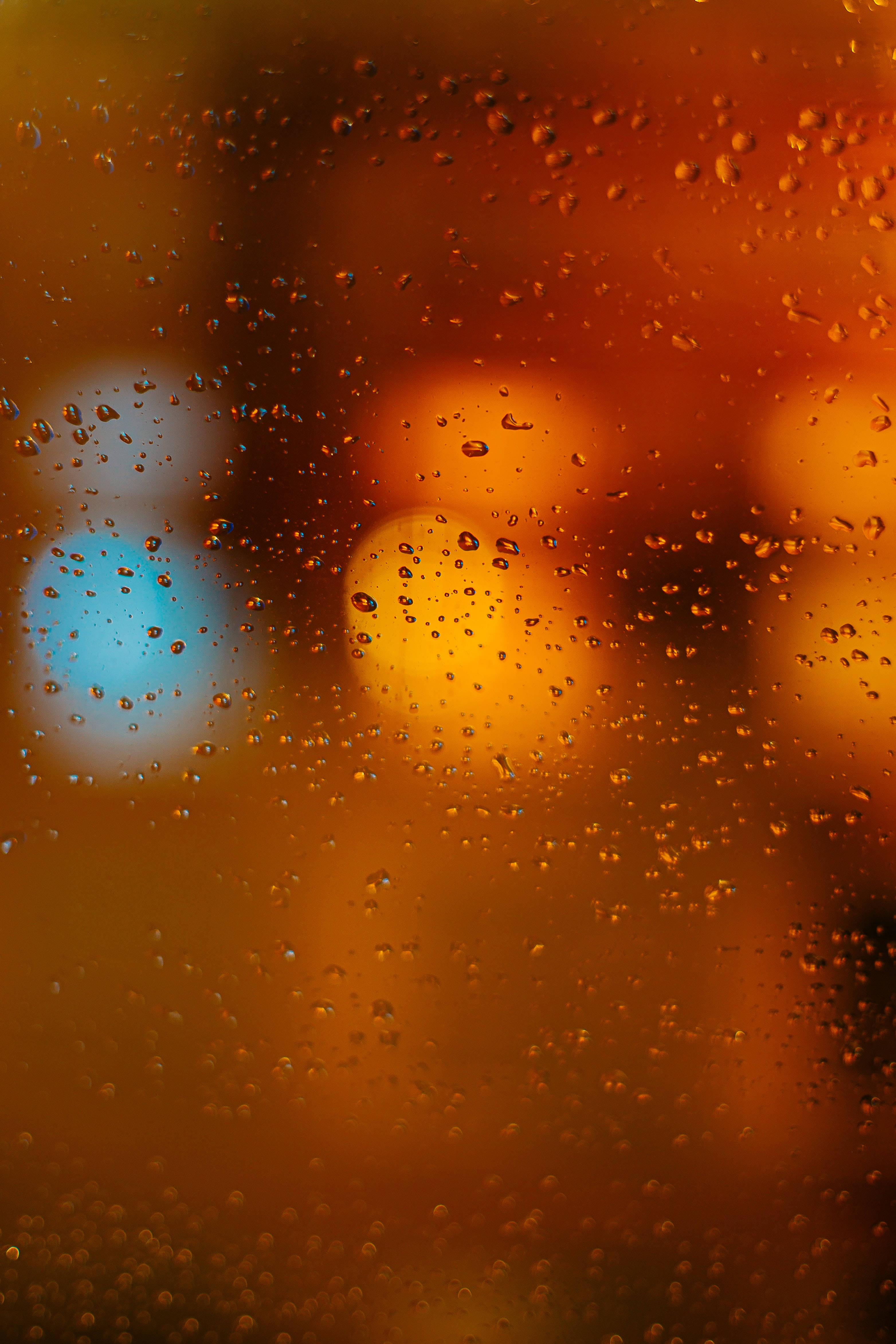 玻璃上的水滴特寫，暖橙色光斑背景 壁紙 — 雨滴依附在窗戶上，顯露出遠處模糊的橙色和藍色城市燈光。