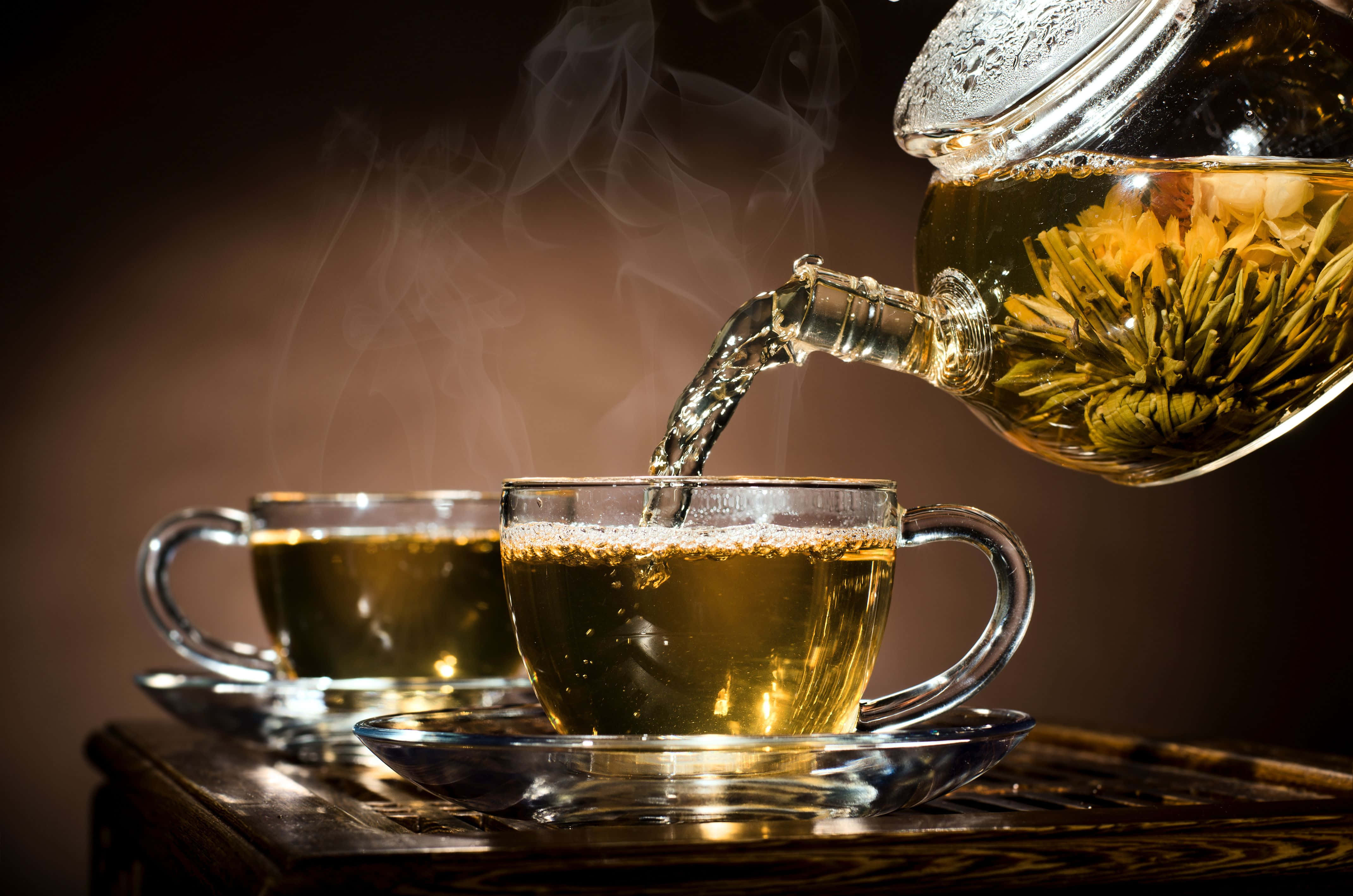 玻璃壺倒茶入杯圖 壁紙 — 冒煙的金色菊花茶從玻璃茶壺倒入木托盤上的透明杯中。