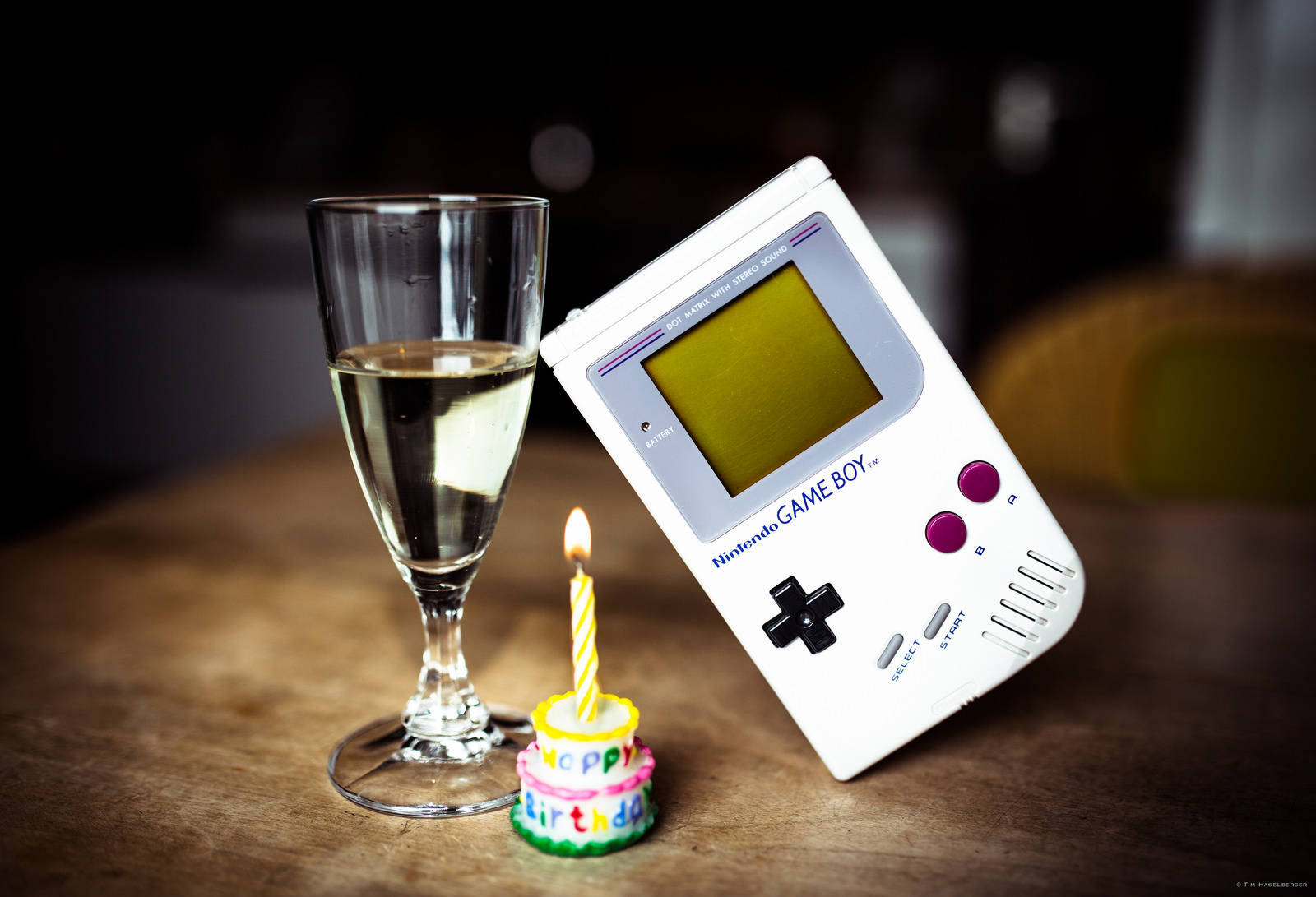 玻璃桌上的復古 Game Boy 與蛋糕 壁紙 — 一部白色 Game Boy 靠喺木枱上嘅香檳杯同插住一支點燃蠟燭嘅細細個生日蛋糕旁邊。