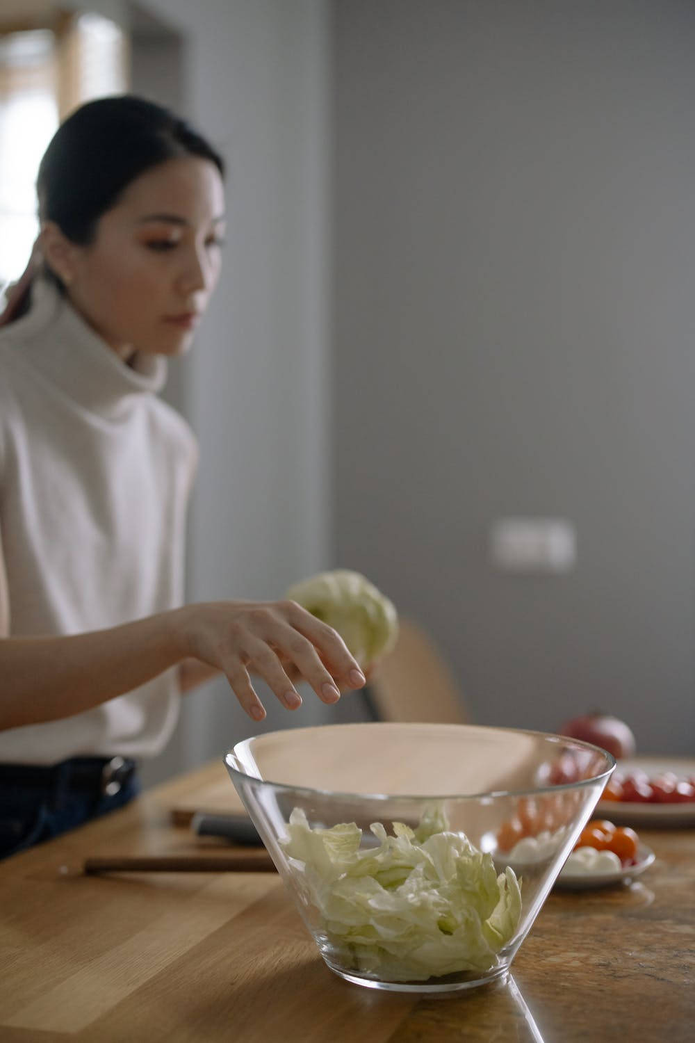 玻璃碗中的捲心菜 壁紙 — 一位身穿白色高領毛衣的女性正在木製廚房檯面上的玻璃碗中準備新鮮沙律。