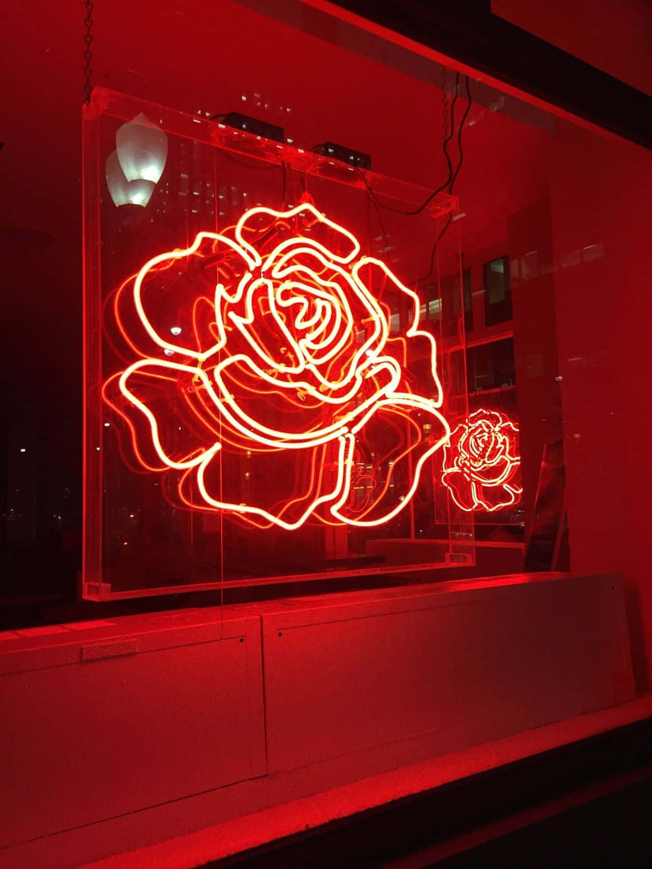 Pastel Red Aesthetic 桌布 壁紙 — 玻璃箱中發光的紅色霓虹玫瑰雕塑，在夜晚向黑暗的城市窗戶投射出溫暖的光芒。