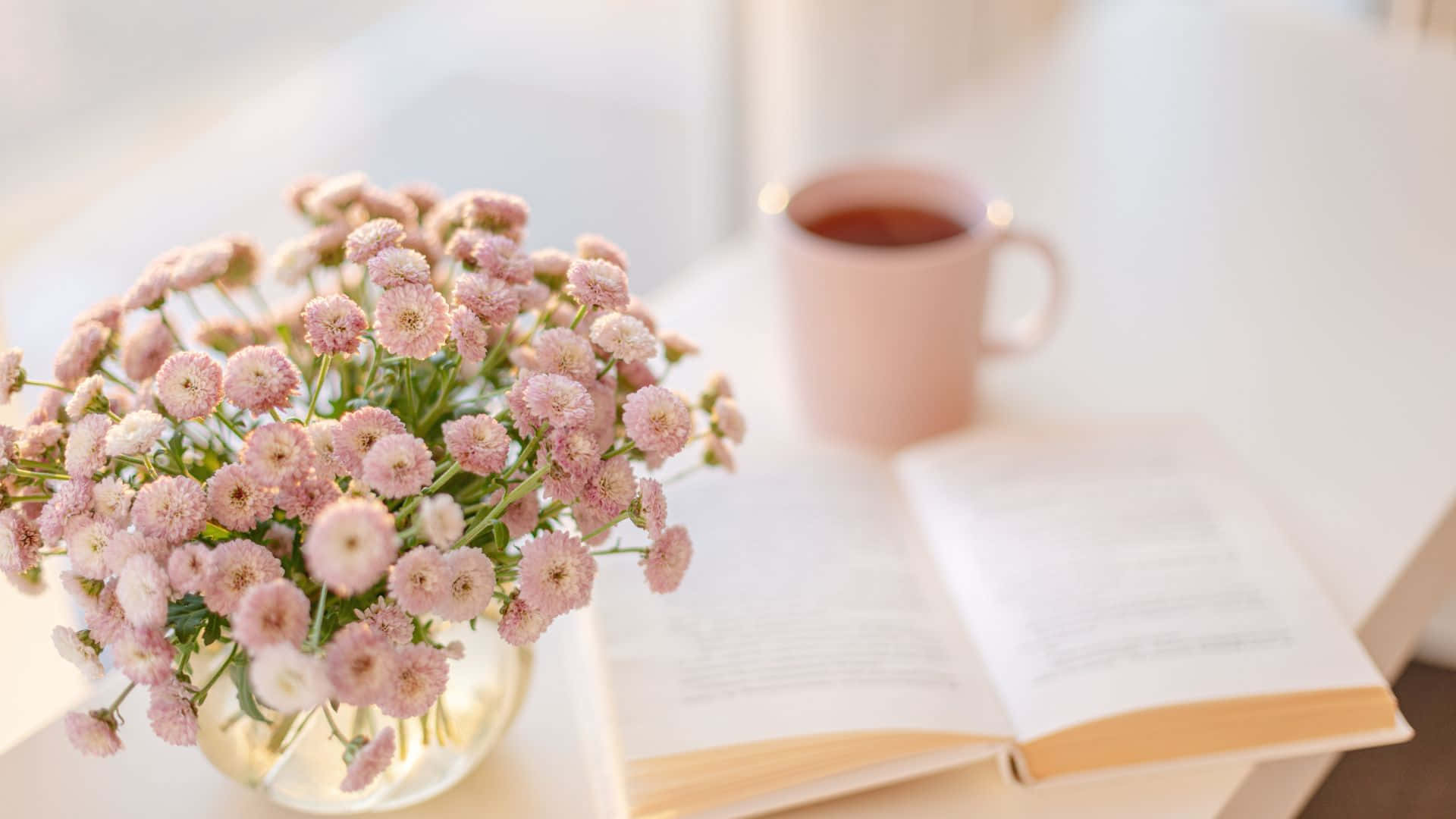 Pink Tea 桌布 壁紙 — 玻璃花瓶中的柔和粉紅色花朵，旁邊放著一本打開的書和一個相配的杯子，在明亮的白色桌子上。