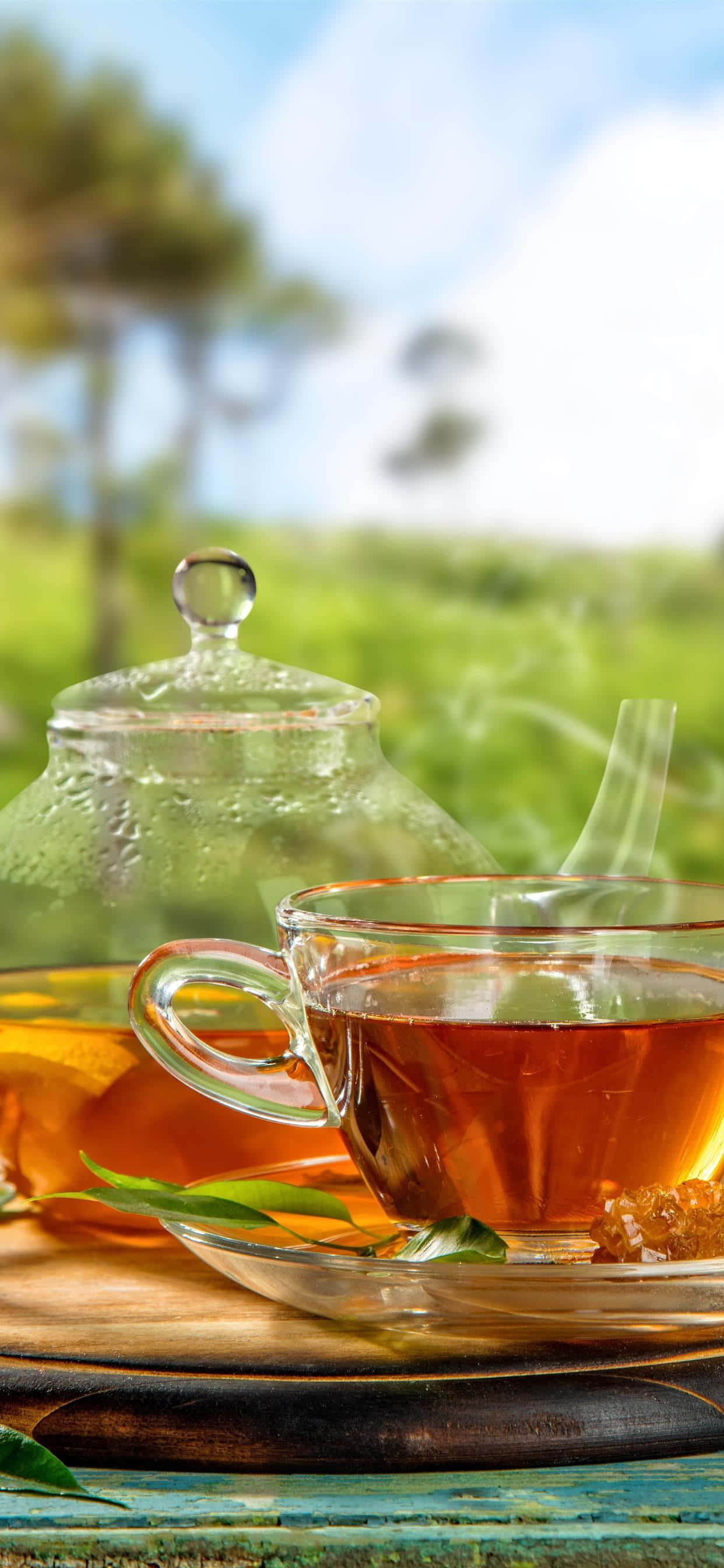 玻璃茶杯與壺圖 壁紙 — 一個冒著蒸氣的玻璃茶杯和茶壺放在戶外一個質樸的木托盤上，背景是明亮的藍天和柔焦的綠樹。