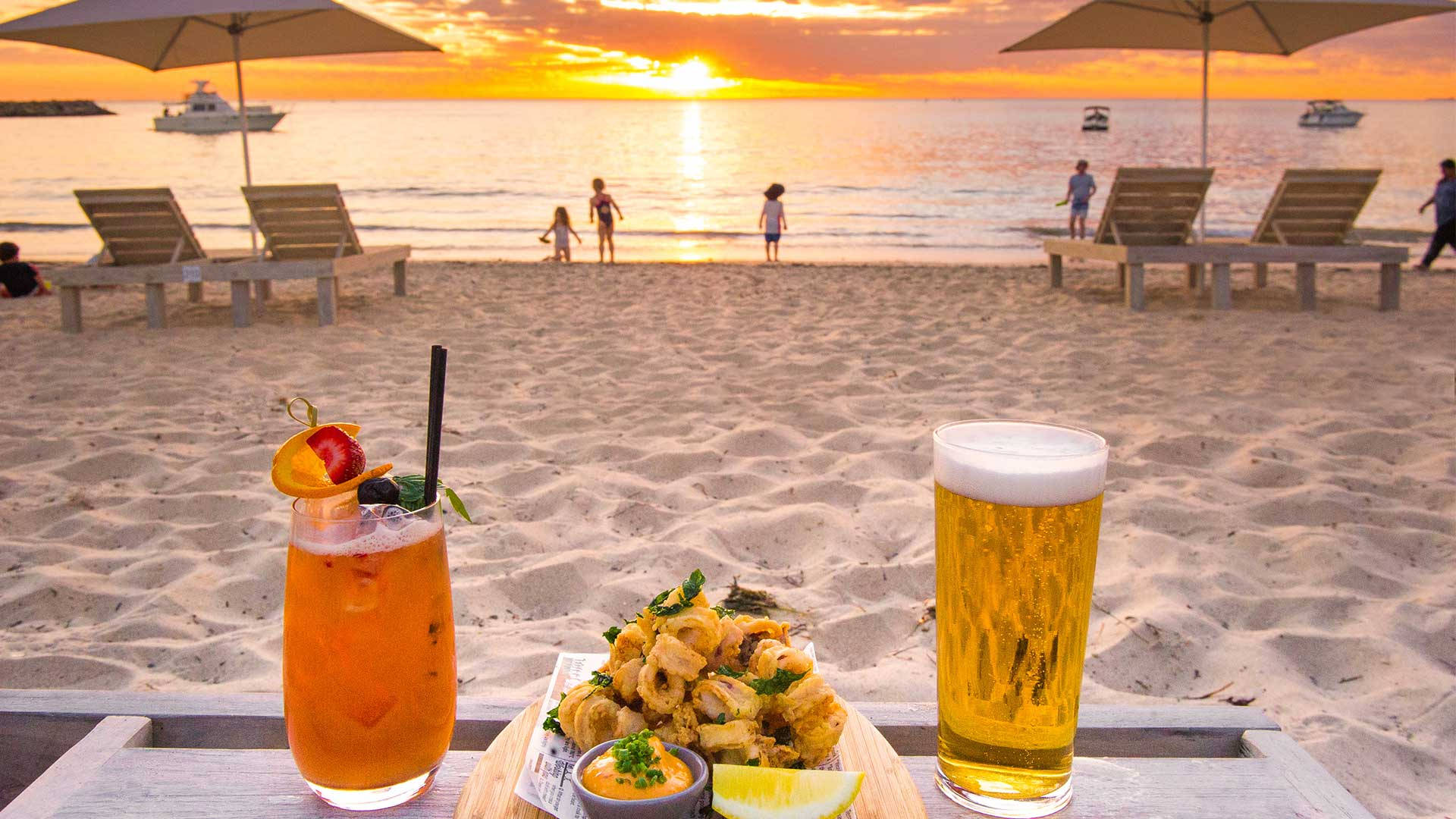 日落海灘場景，桌子上有飲料和小吃，孩子們在水中玩耍。