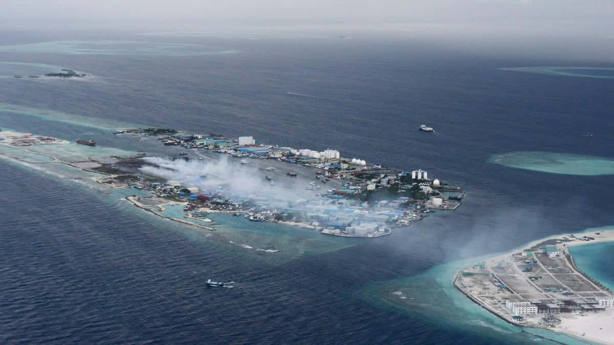 Maldives Island 桌布 壁紙 — 珊瑚環礁島嶼的鳥瞰圖，在碧綠潟湖和深藍色海洋中，火災產生的煙霧升騰。