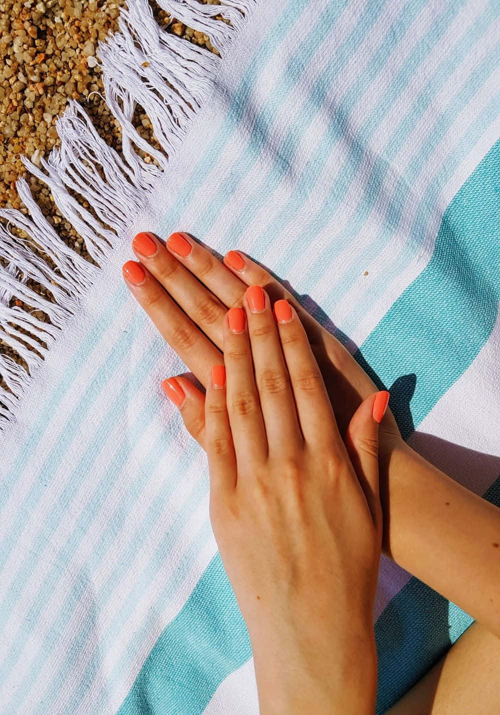 Cute Nails 桌布 壁紙 — 珊瑚色指甲的手在明亮陽光下，擱在卵石海灘旁的藍白條紋沙灘巾上。