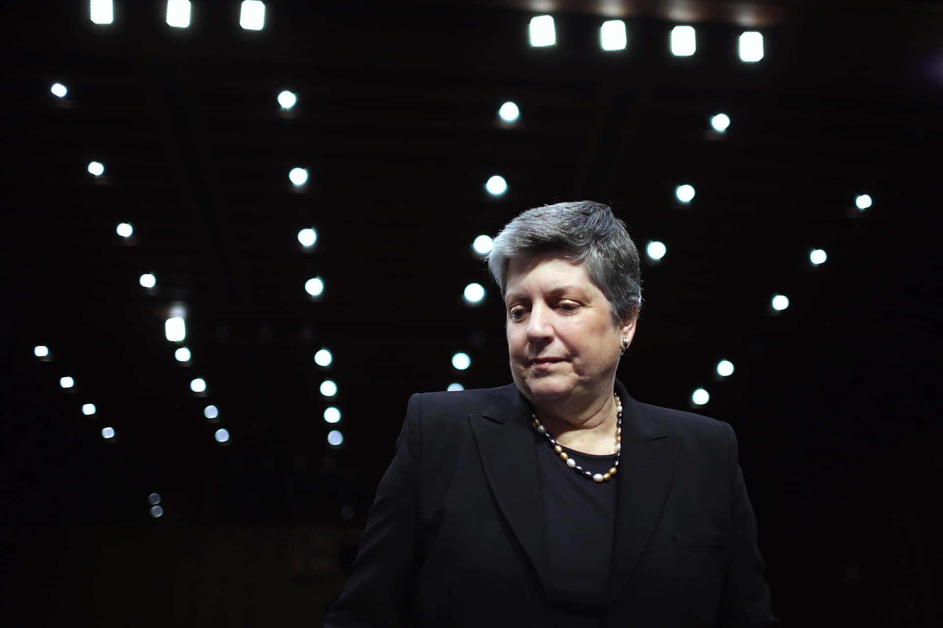 一位穿著黑色西裝和珍珠項鍊的女士站在舞台燈光下。