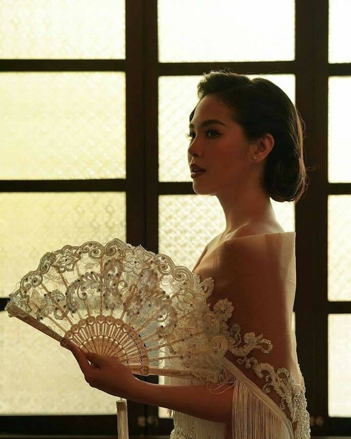 身穿白色蕾絲長裙的女性，在金色光線的窗邊手持華麗扇子。