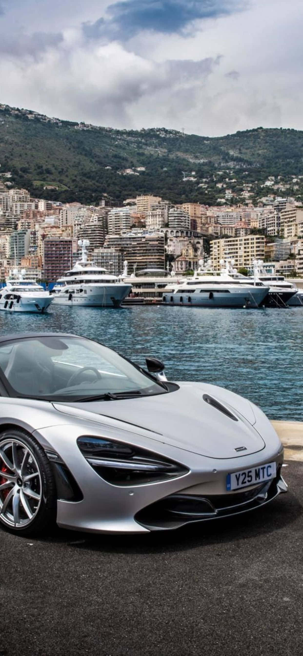 珍珠白 iPhone Xs Max McLaren 720s 背景 壁紙 — 一架時尚的銀色跑車停泊在海濱，背景有豪華遊艇和山丘城市。