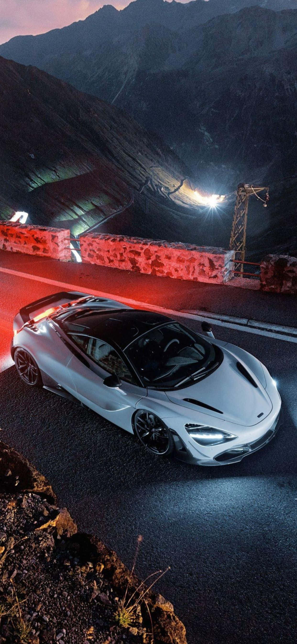 珍珠白 iPhone Xs Max McLaren 720s 背景 壁紙 — 一輛時尚的銀色跑車在黃昏時分沿著蜿蜒的山路行駛，車頭燈閃爍，周圍環繞著紅色燈光。