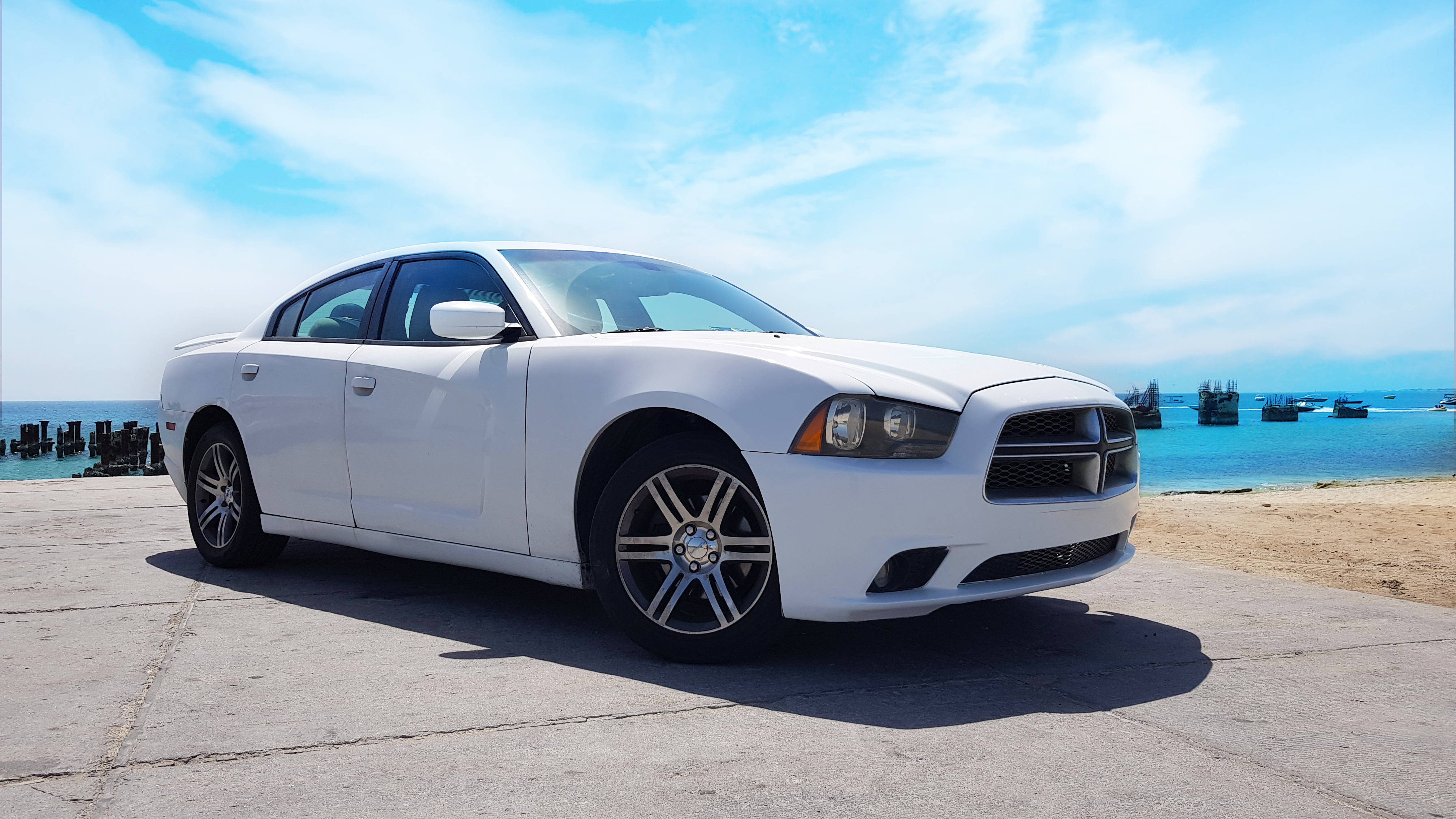 珍珠白塗裝的道奇挑戰者 壁紙 — 一輛純白色的 Dodge Charger 停泊在混凝土碼頭上，碧綠色的海洋和遠處的船隻在明亮的藍天下。