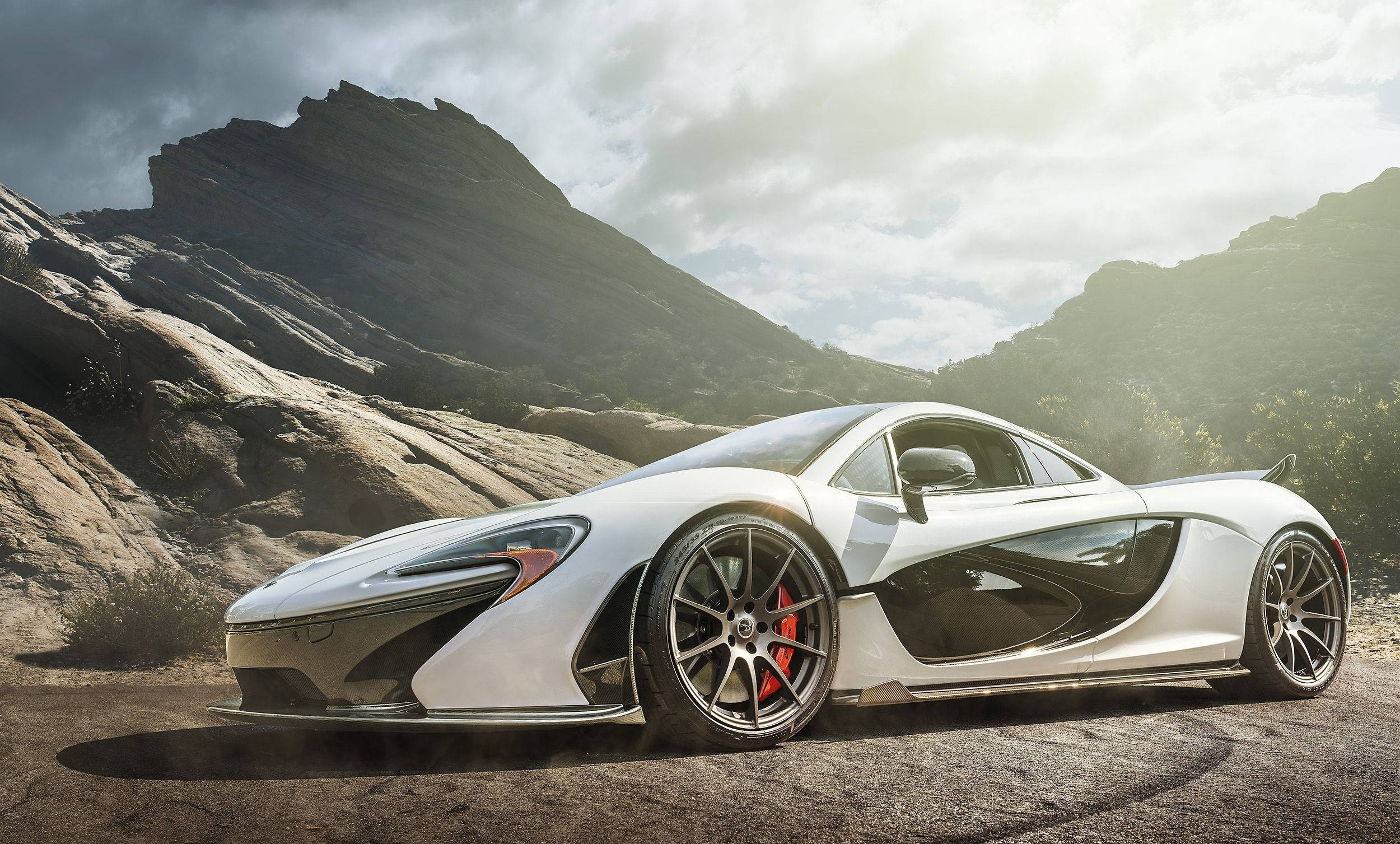珍珠白麥拉倫P1 壁紙 — 一架時尚的白色McLaren跑車，帶有紅色制動卡鉗，停泊在一條塵土飛揚的道路上，背景是戲劇性的岩石山脈，天空多雲。