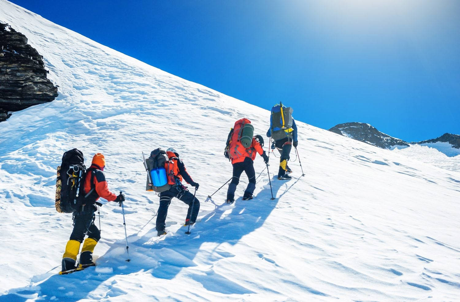 四名登山者穿著鮮豔裝備在藍天下攀登雪山斜坡。
