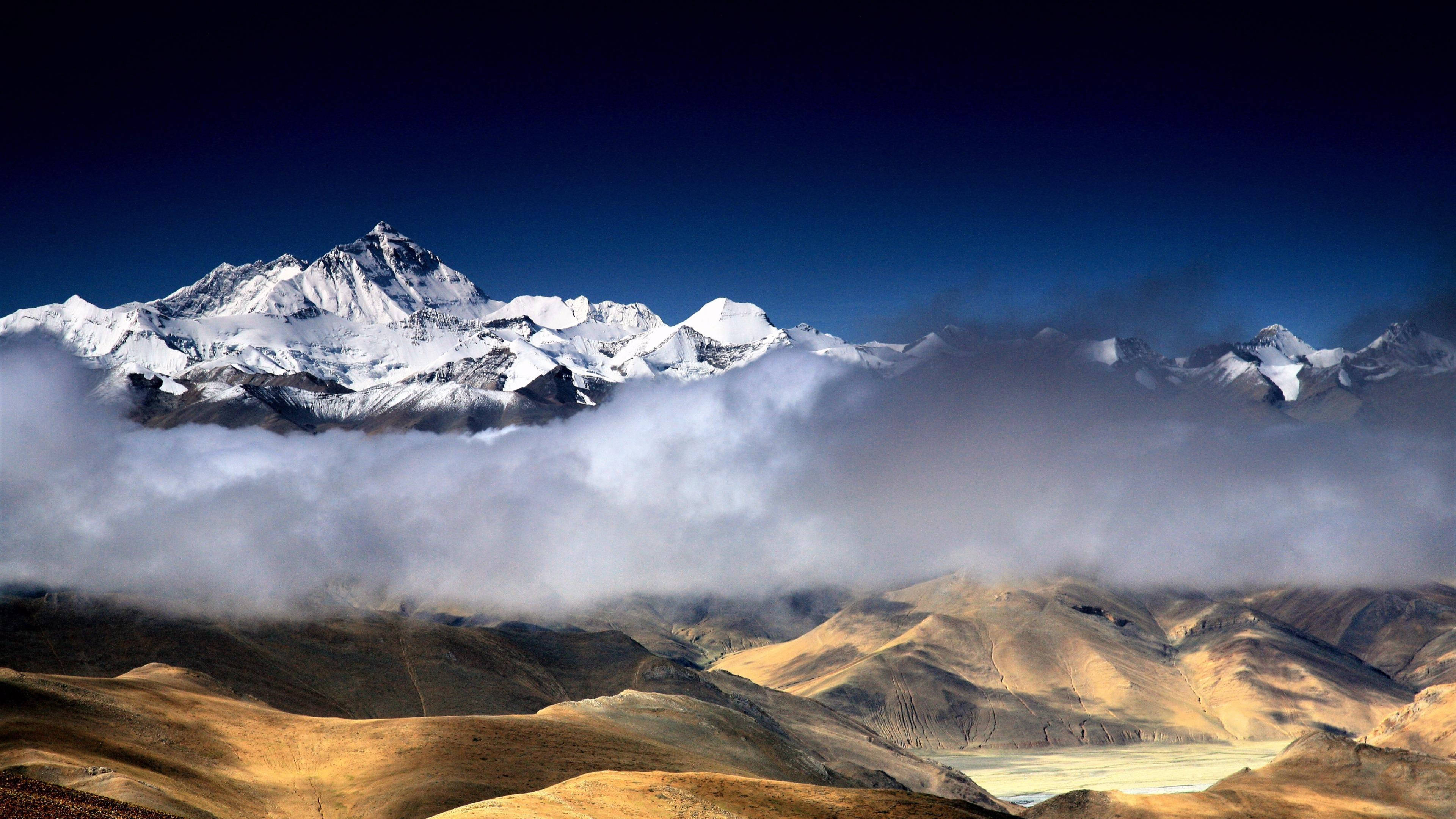 珠峰壯麗全景 壁紙 — 白雪覆蓋的山峰穿過厚厚的雲層，在深藍色的天空下，金黃色的乾旱山谷之上。