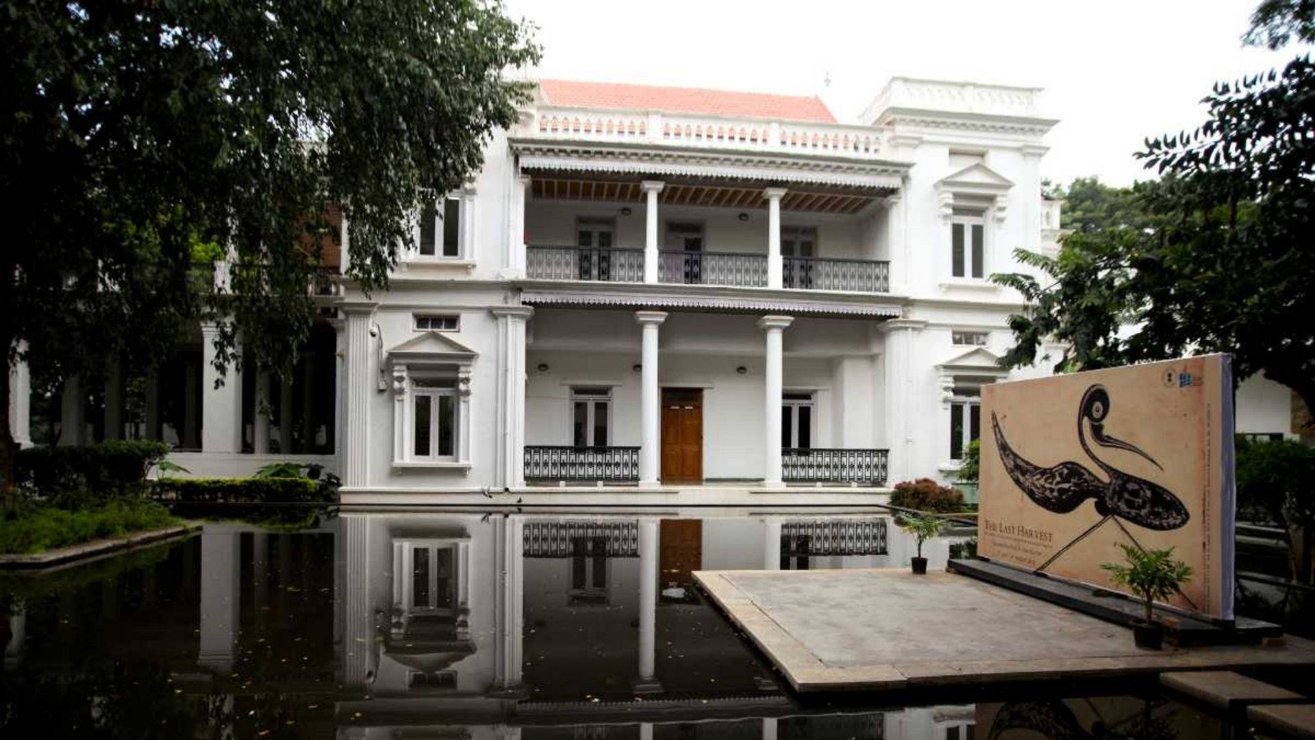 班加羅爾國家美術館：藝術遺產一瞥 壁紙 — 一座宏偉的白色殖民風格大宅，帶有柱子和陽台，完美倒映在平靜的池塘中，周圍環繞著茂密的綠樹。
