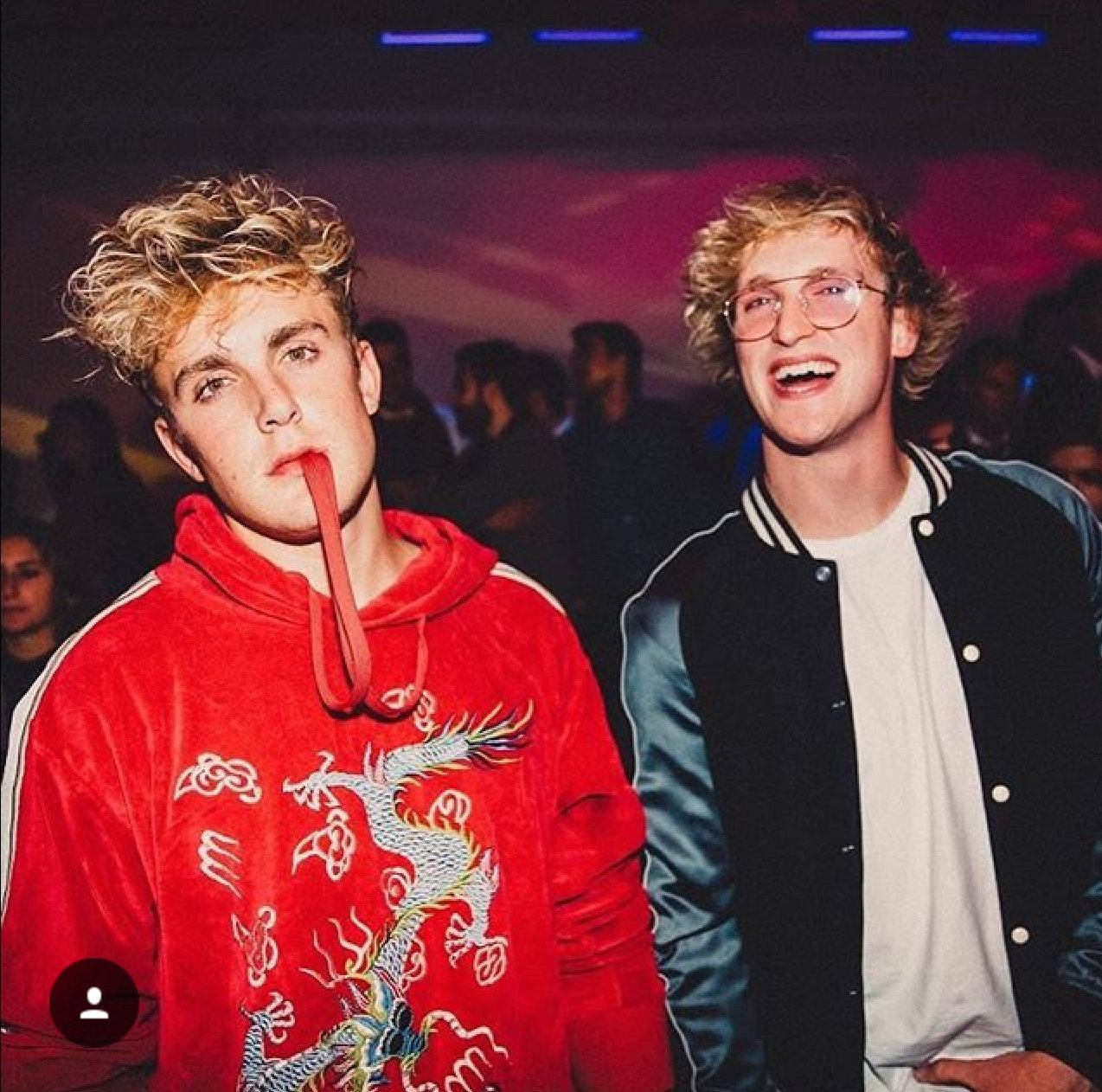 現代 Maverick - Logan Paul 動感時刻 壁紙 — 兩個年輕男子，一頭金色捲髮，在燈光昏暗的派對上擺姿勢，一個穿著紅色龍圖案連帽衫，另一個穿著棒球外套，笑著。