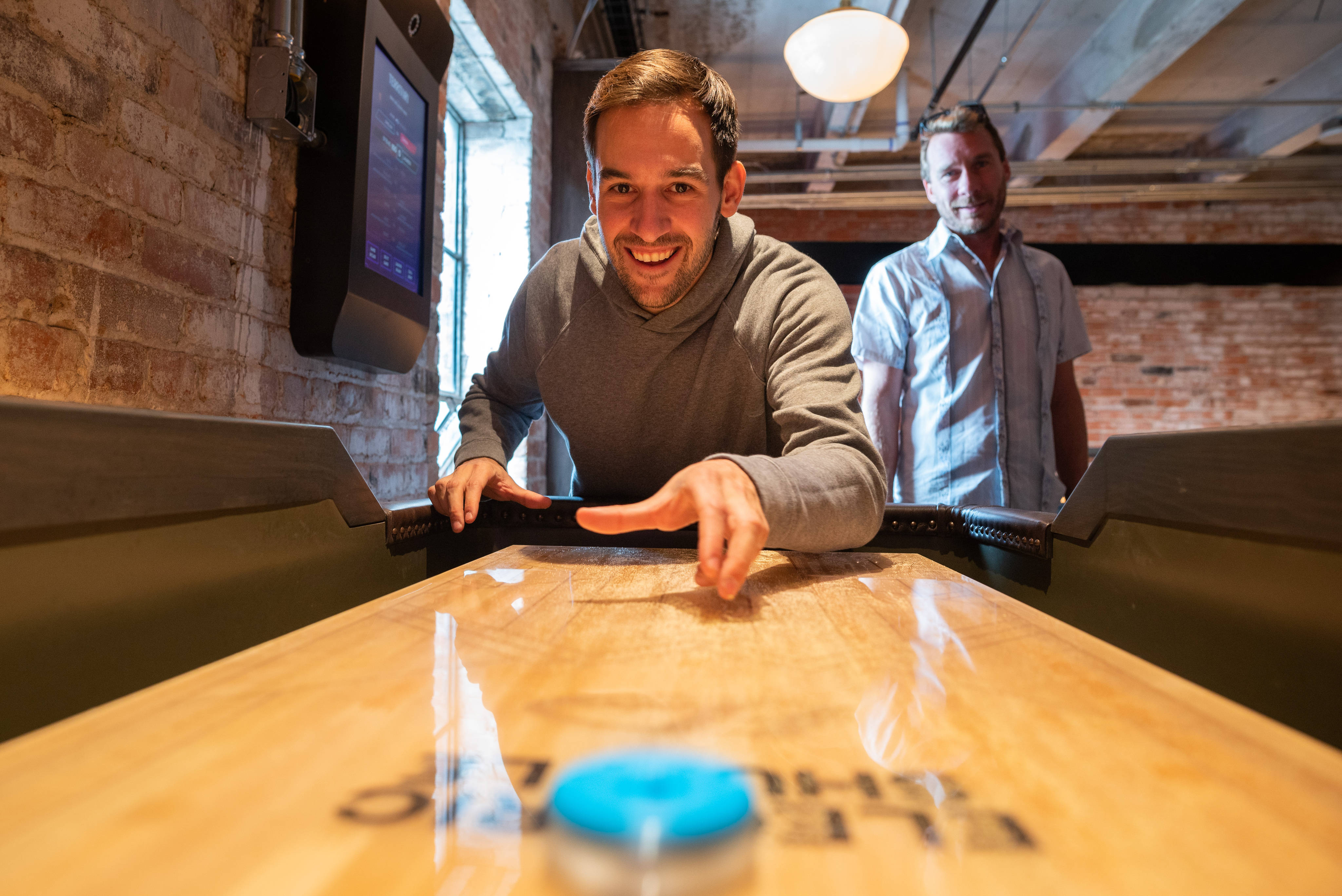 現代 Shuffleboard 桌與藍色球 壁紙 — 一個微笑的男人靠在一張沙狐球桌上，焦點在一個藍色冰球上，另一個男人在背景中看著，背景是裸露的磚牆。